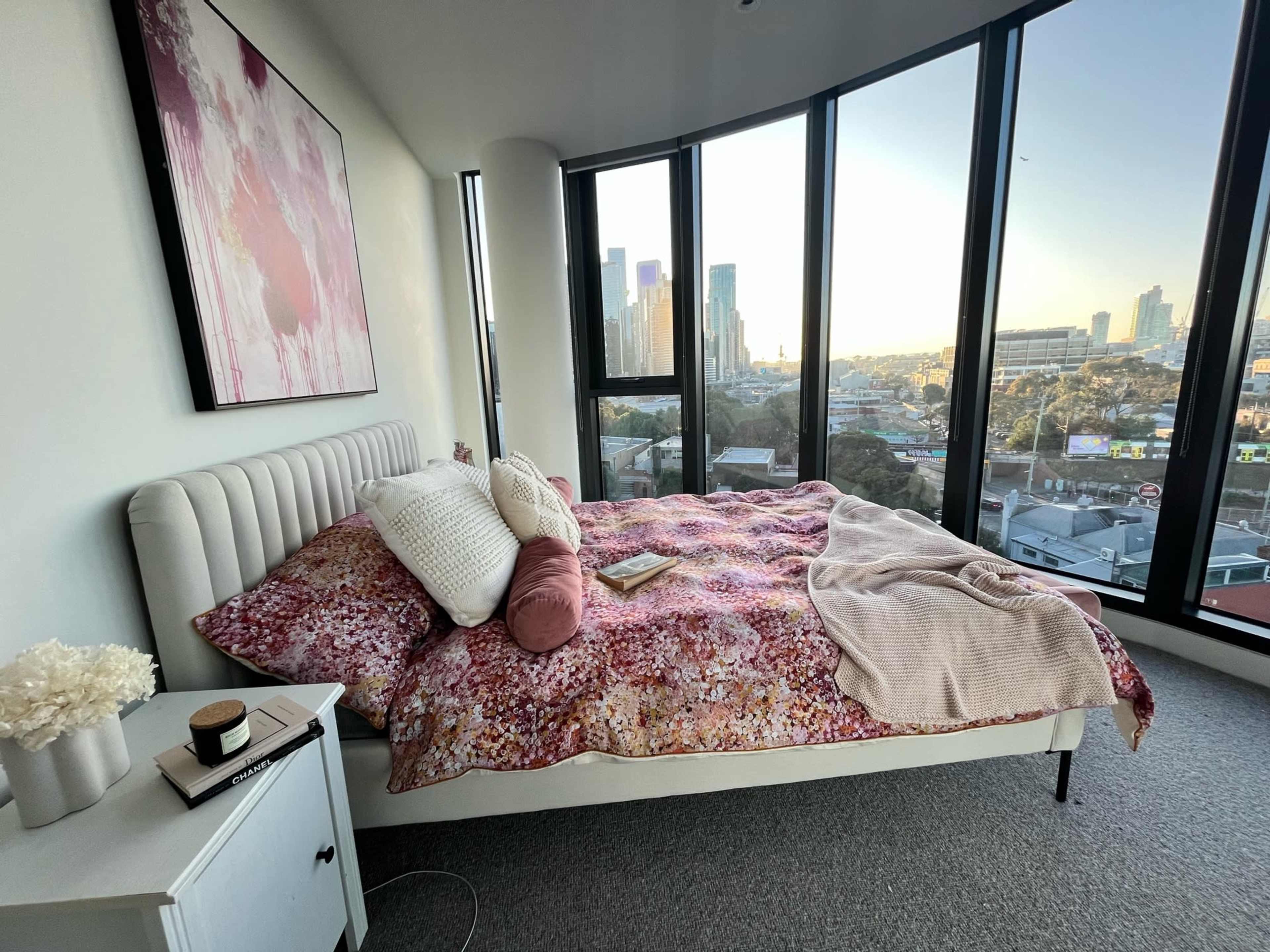 A modern bedroom features a bed with a colorful patterned duvet and a view of the city skyline through large windows.