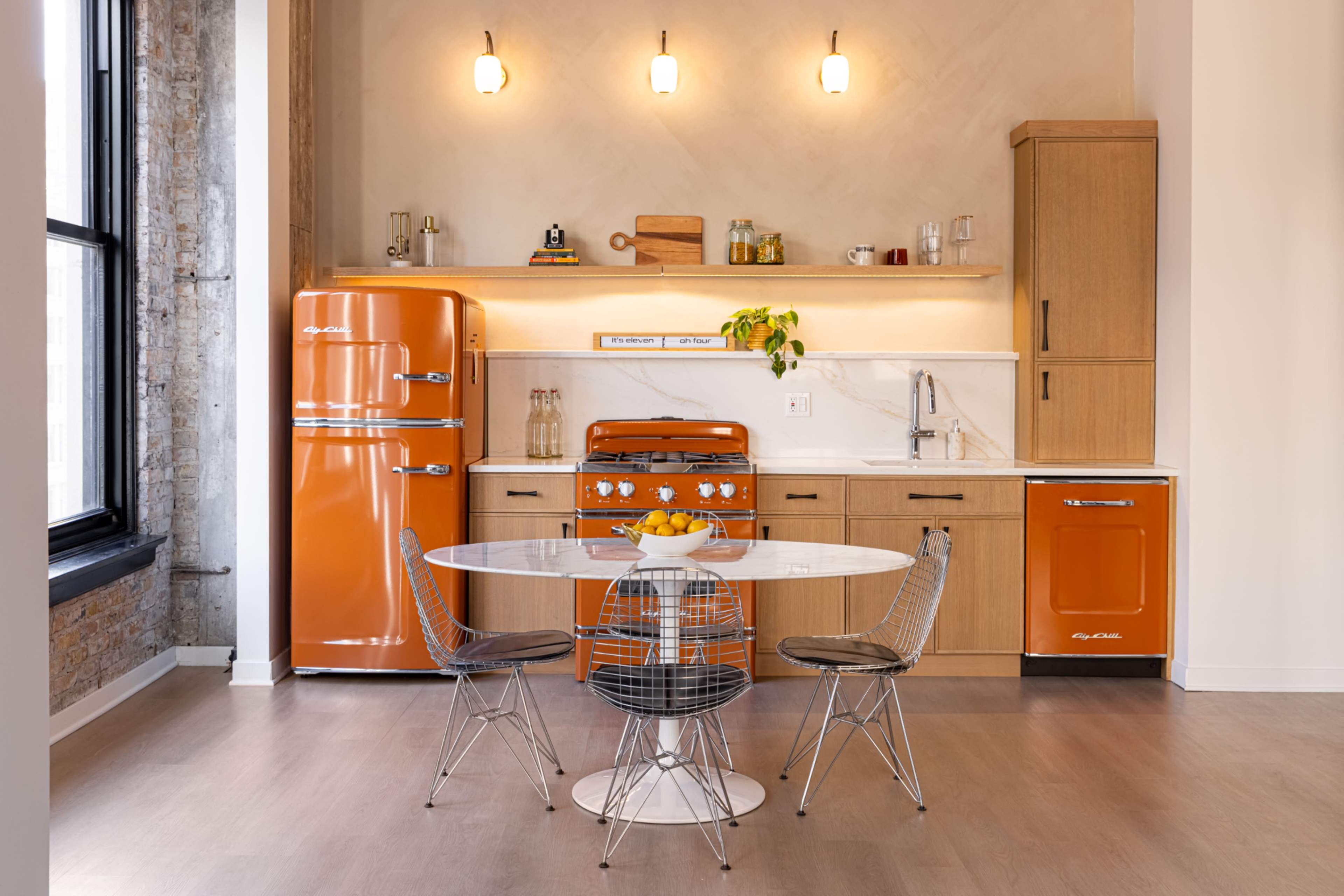 The image shows a modern kitchen with orange appliances, wooden cabinetry, a round dining table, and three wireframe chairs under pendant lighting.