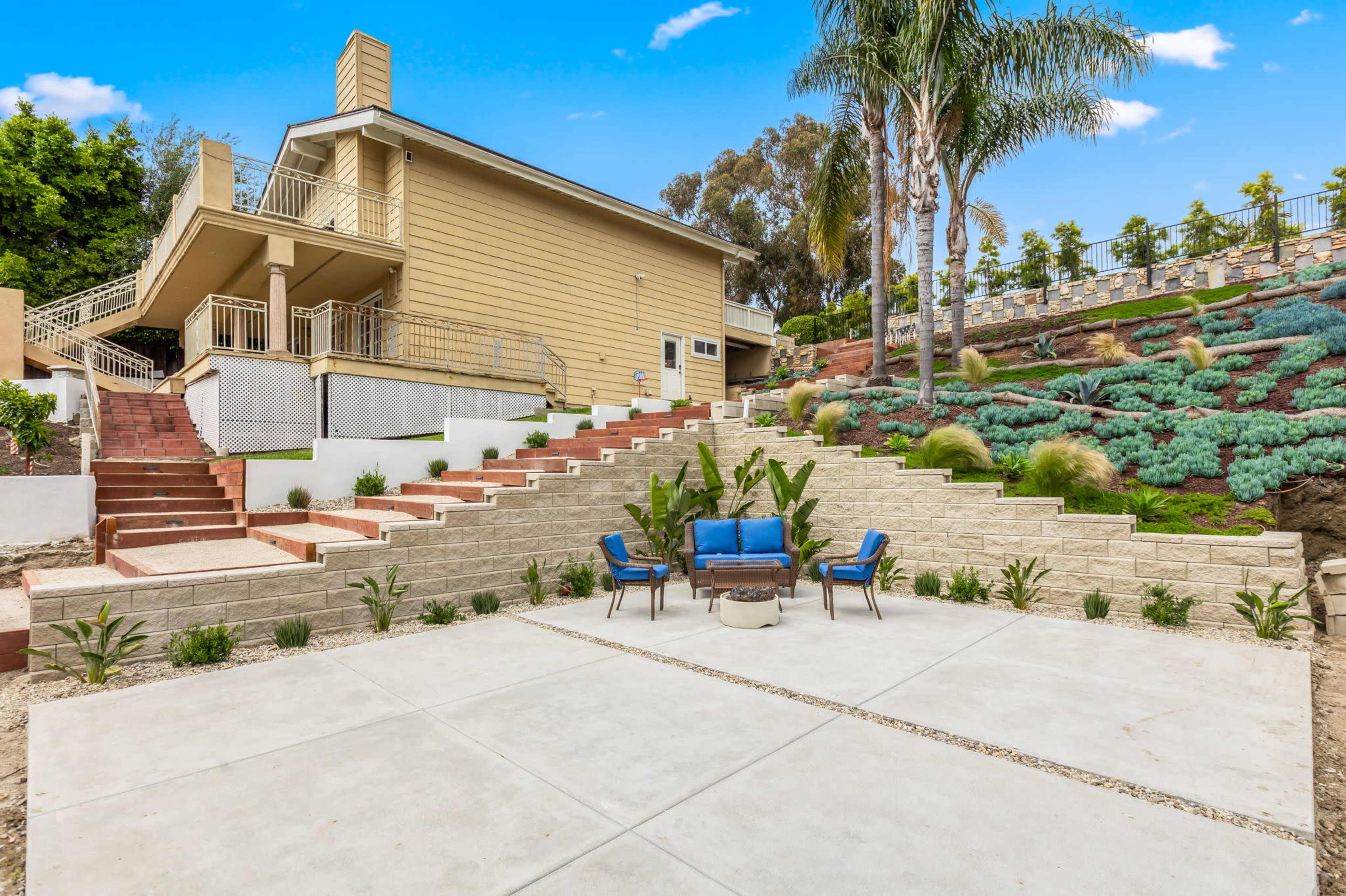 A multi-level outdoor patio features a seating area with blue chairs and a small table, surrounded by landscaped steps and greenery near a large house.