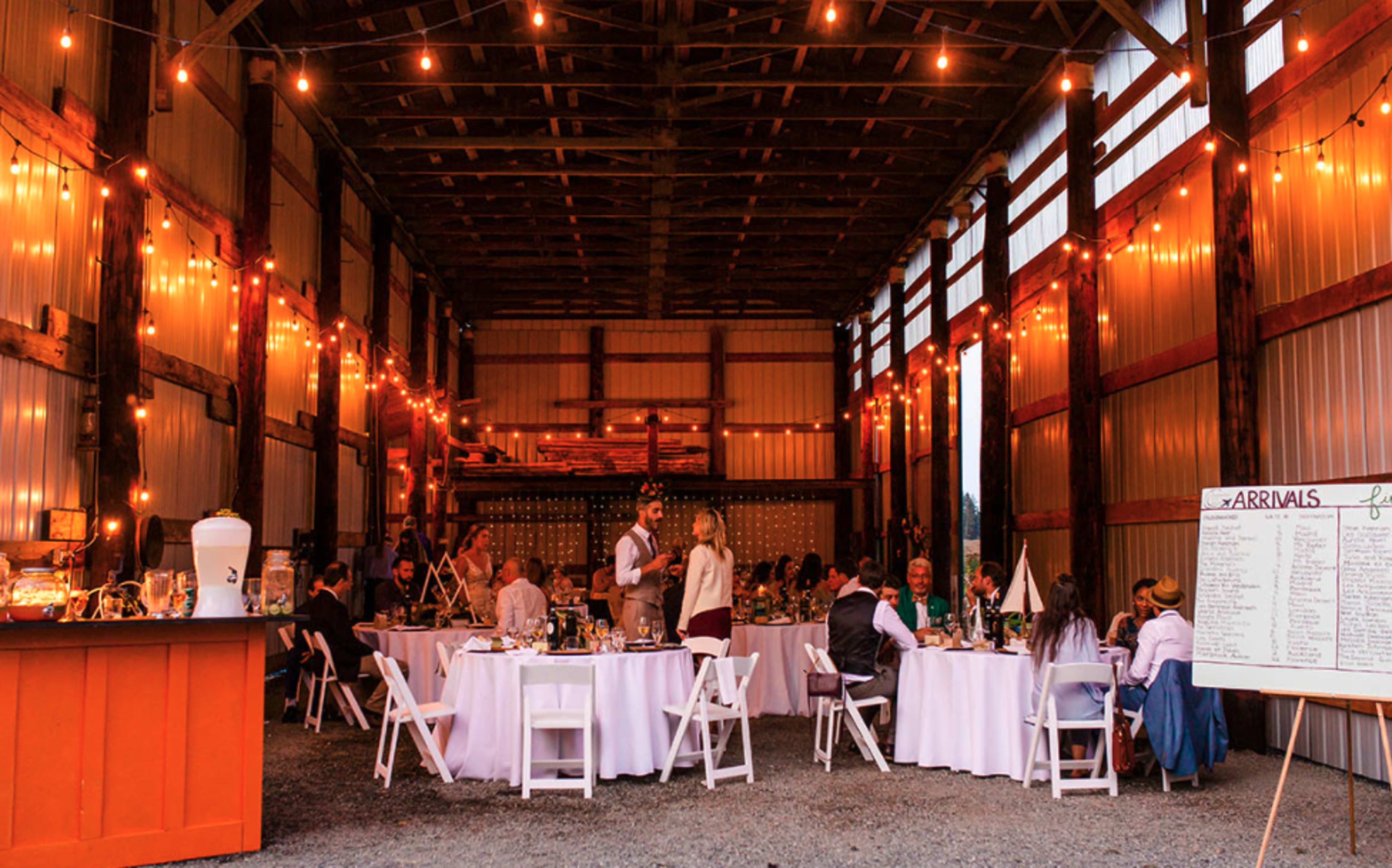 A decorated barn interior with tables set for a wedding reception, string lights illuminating the space, and guests seated at tables.