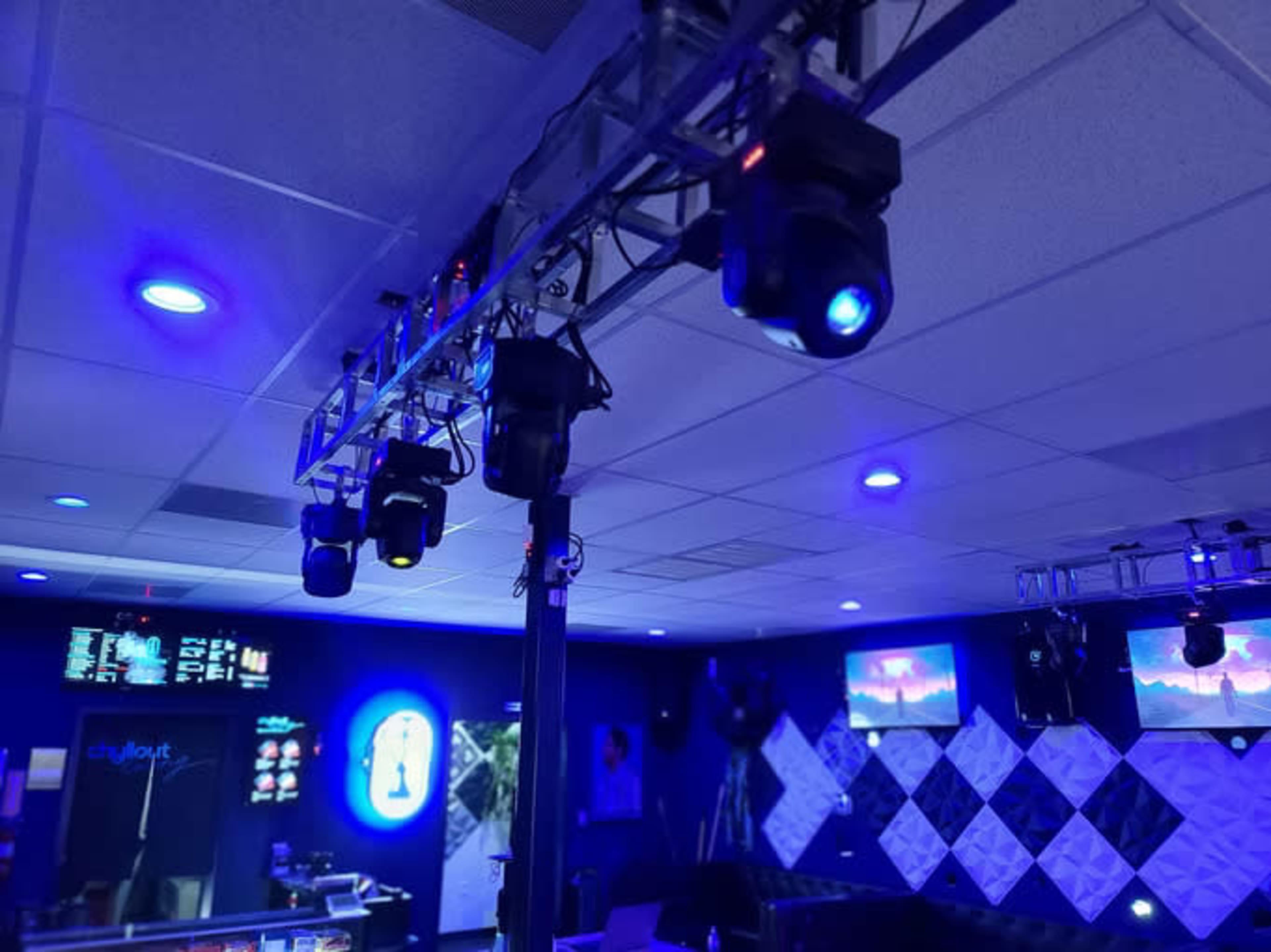 The image shows a dimly lit room equipped with a lighting truss and various stage lights, along with screens displaying information on the walls.