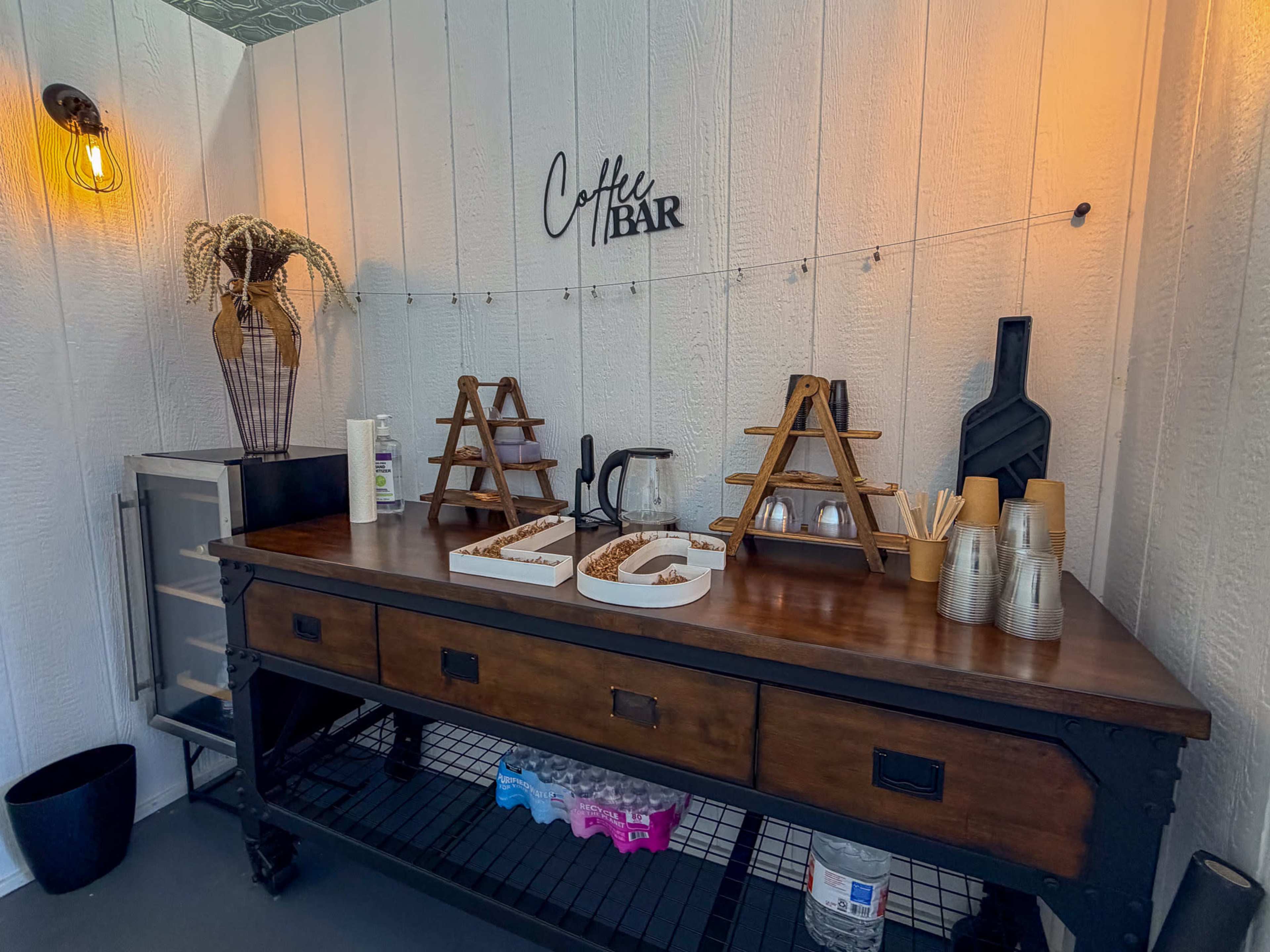 The image shows a rustic coffee bar setup featuring a wooden table with various coffee supplies, including containers for drinks, utensils, and decorative elements on the wall.