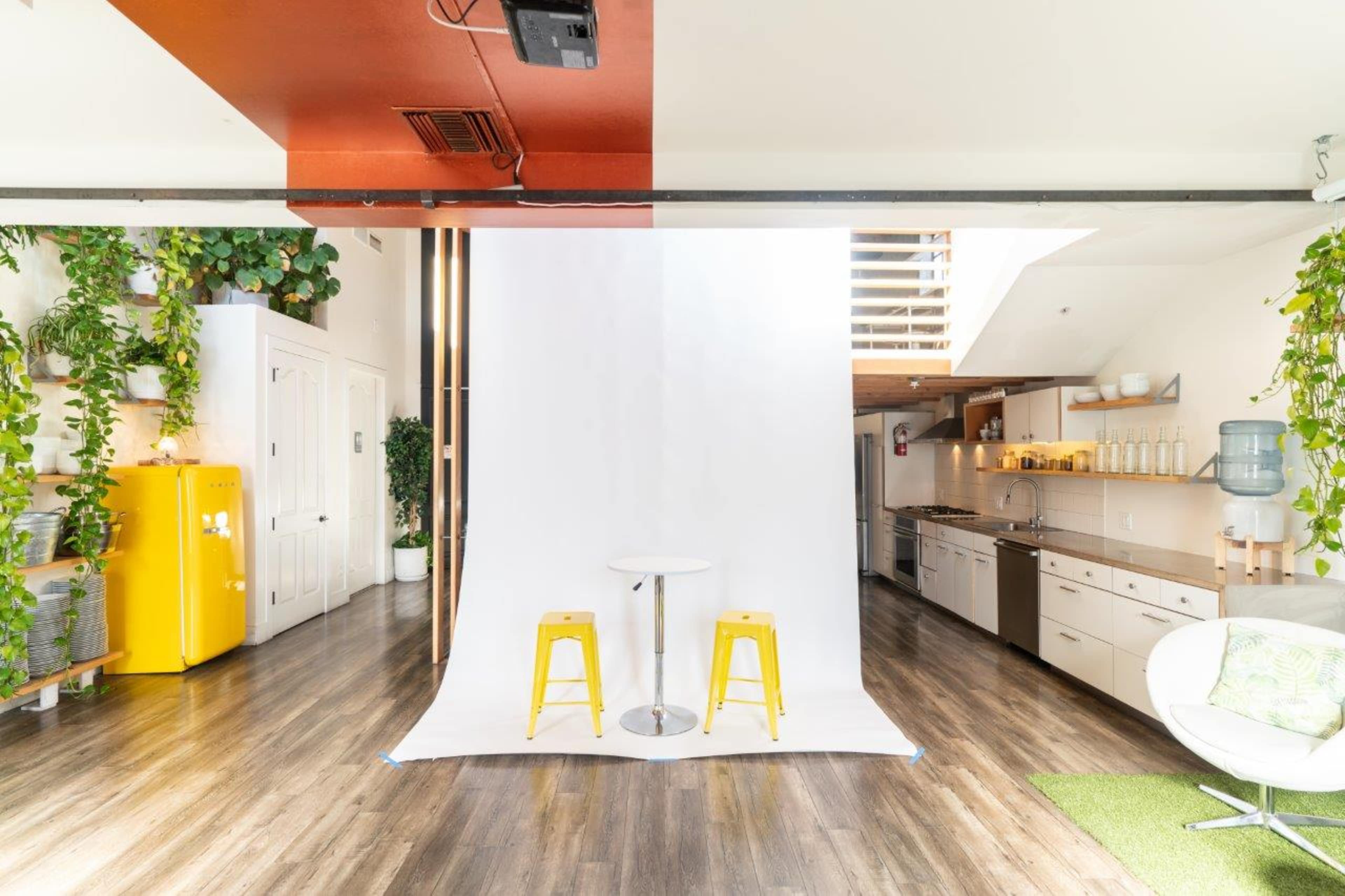 A bright room with a white backdrop and two yellow stools set in front of a kitchen area adorned with plants and a yellow refrigerator.