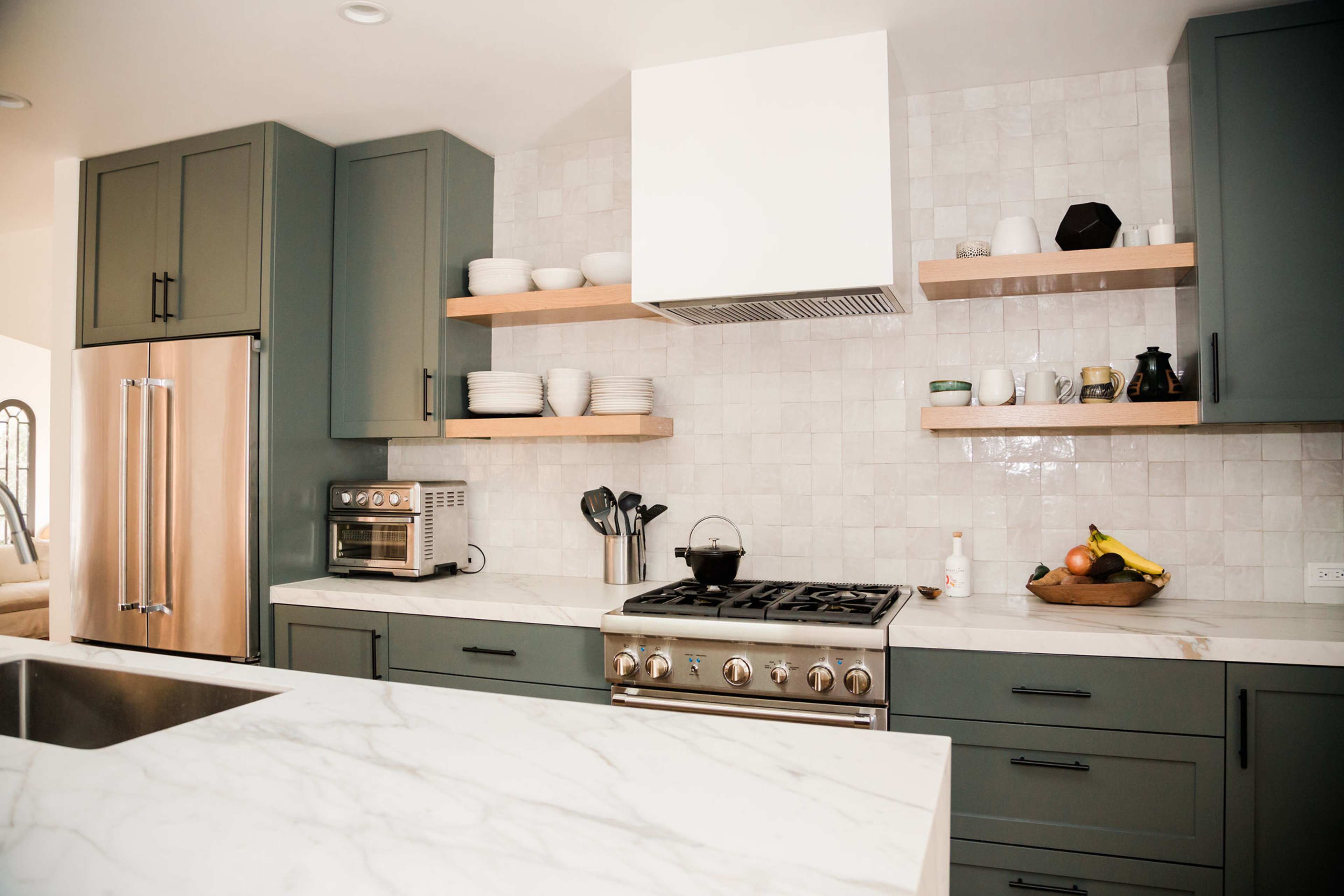 A modern kitchen features green cabinets, a stainless steel refrigerator, and an island with a marble countertop, accompanied by open shelves displaying dishware.
