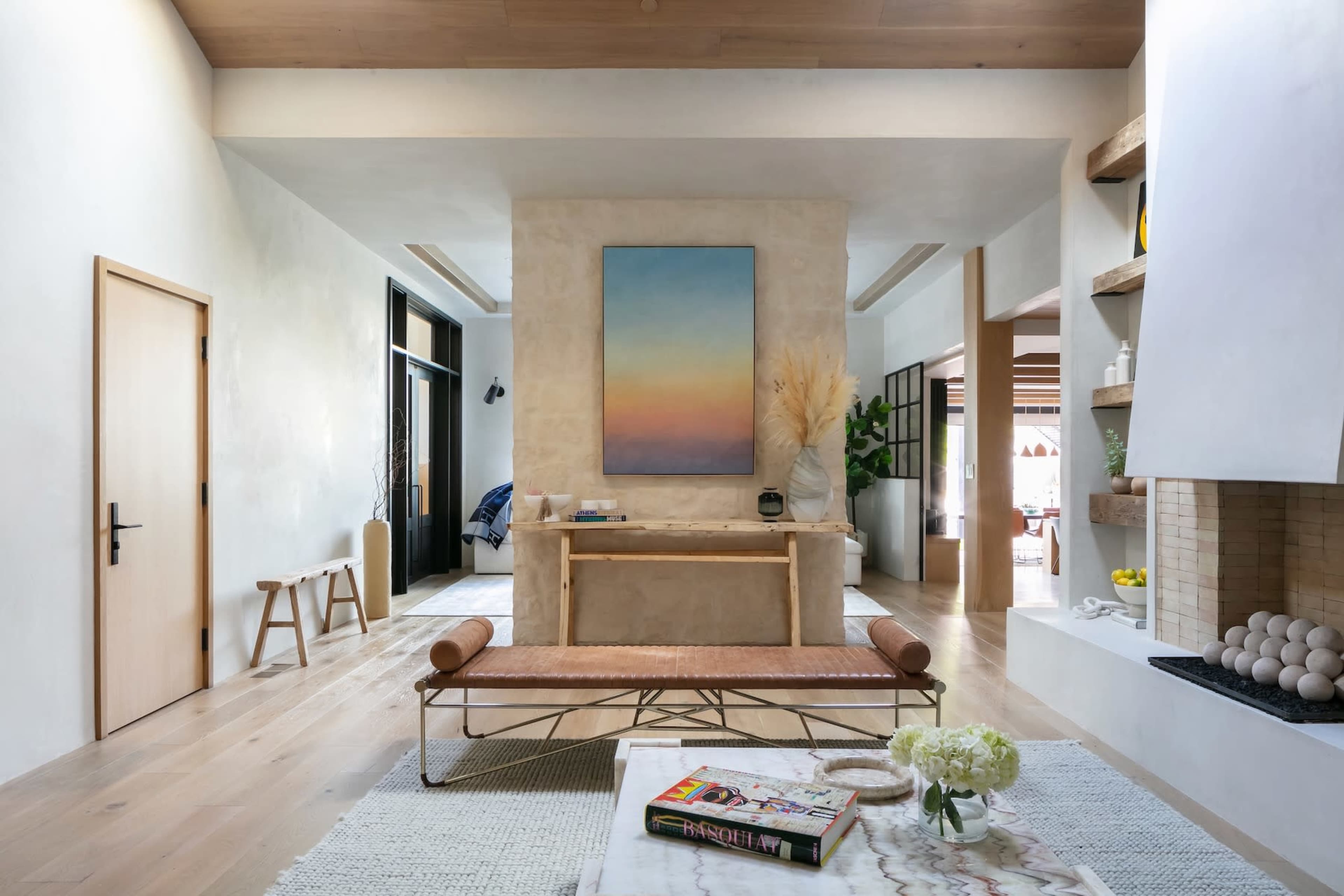 A modern living space featuring a minimalist design with wooden flooring, a neutral color palette, and decorative elements like a large painting and a coffee table.