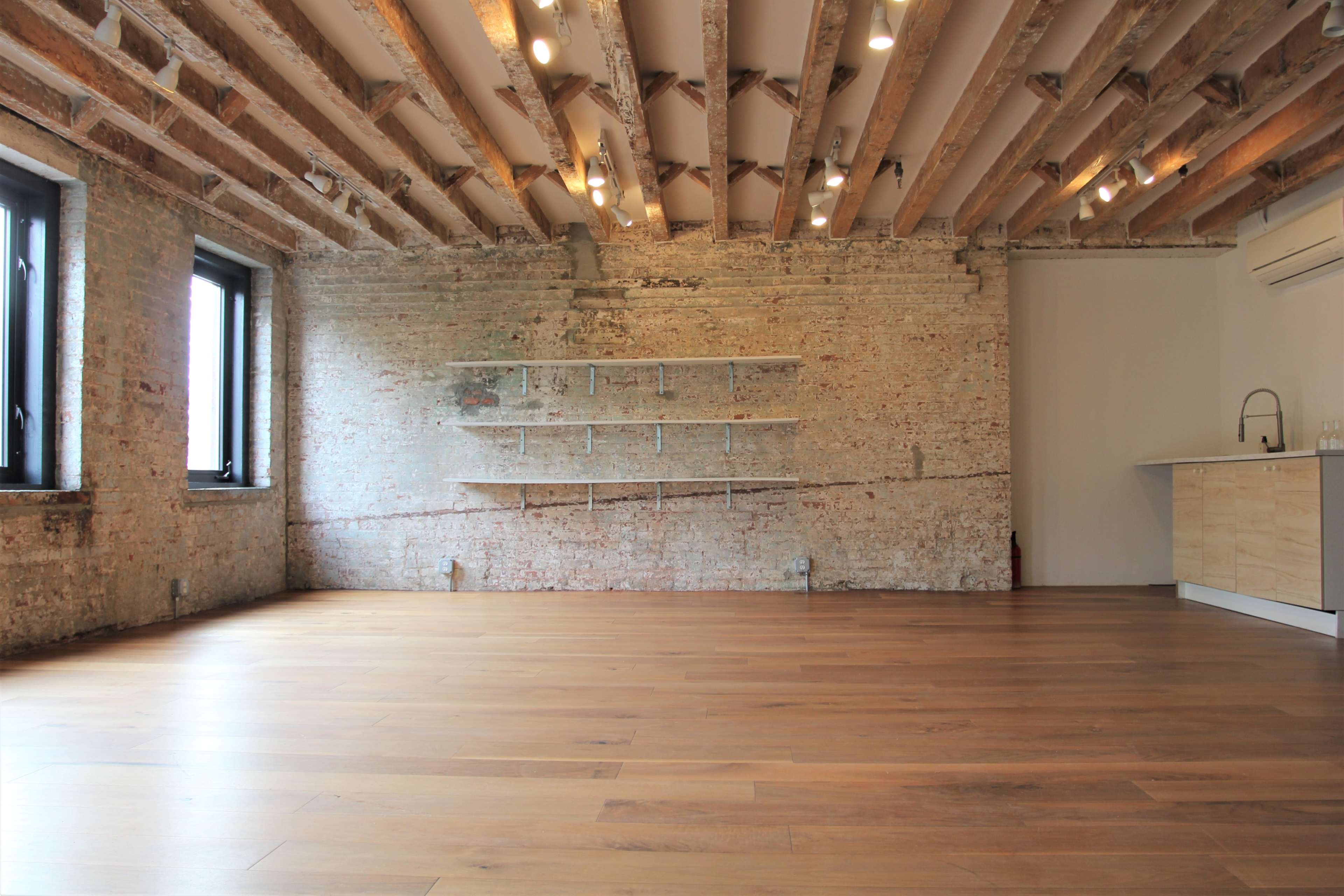 A spacious room with exposed wooden beams, brick walls, and a wooden floor, featuring a simple wall shelf and a kitchenette in one corner.