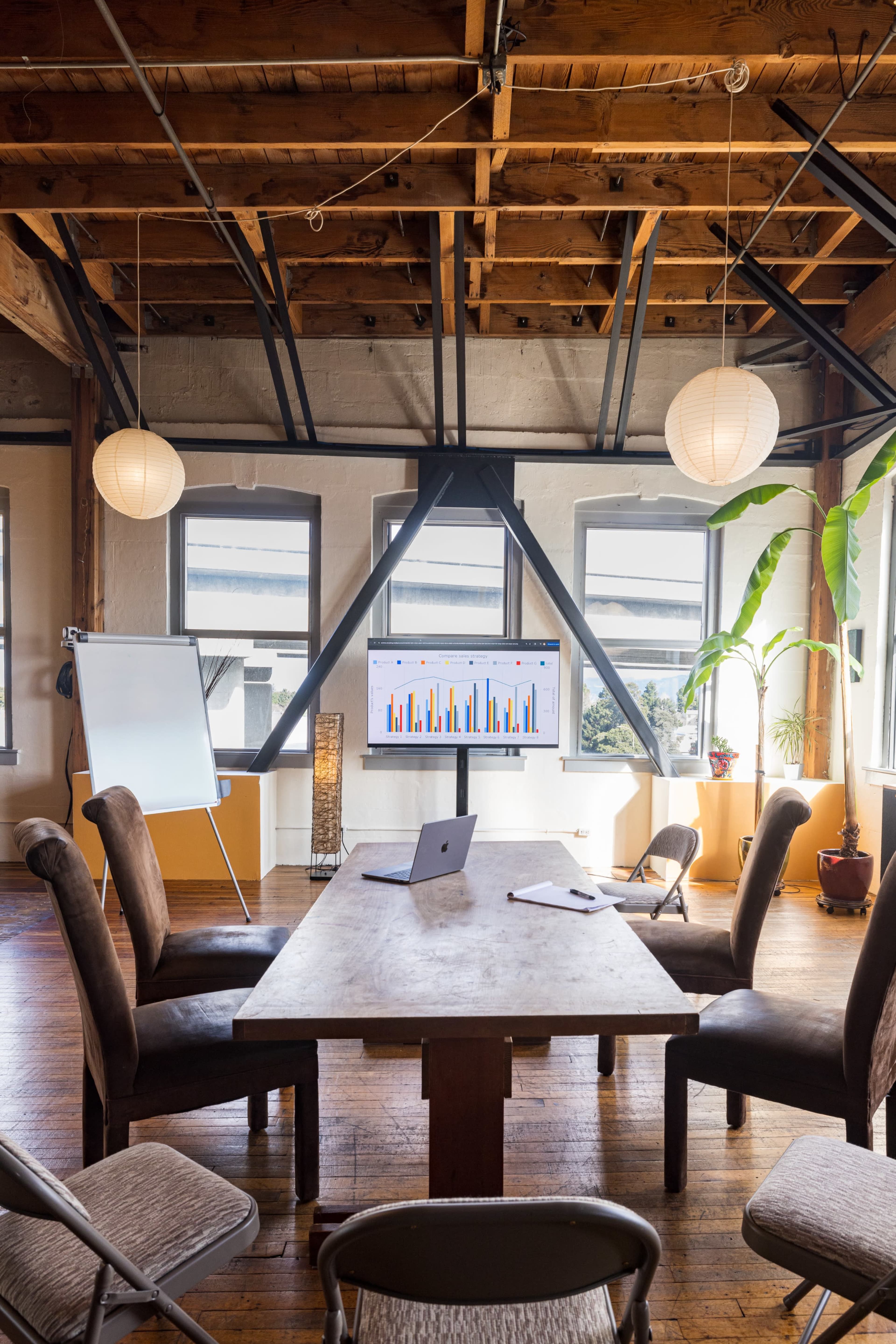 A conference room features a wooden table surrounded by chairs, with a screen displaying colorful bar graphs and large windows letting in natural light.