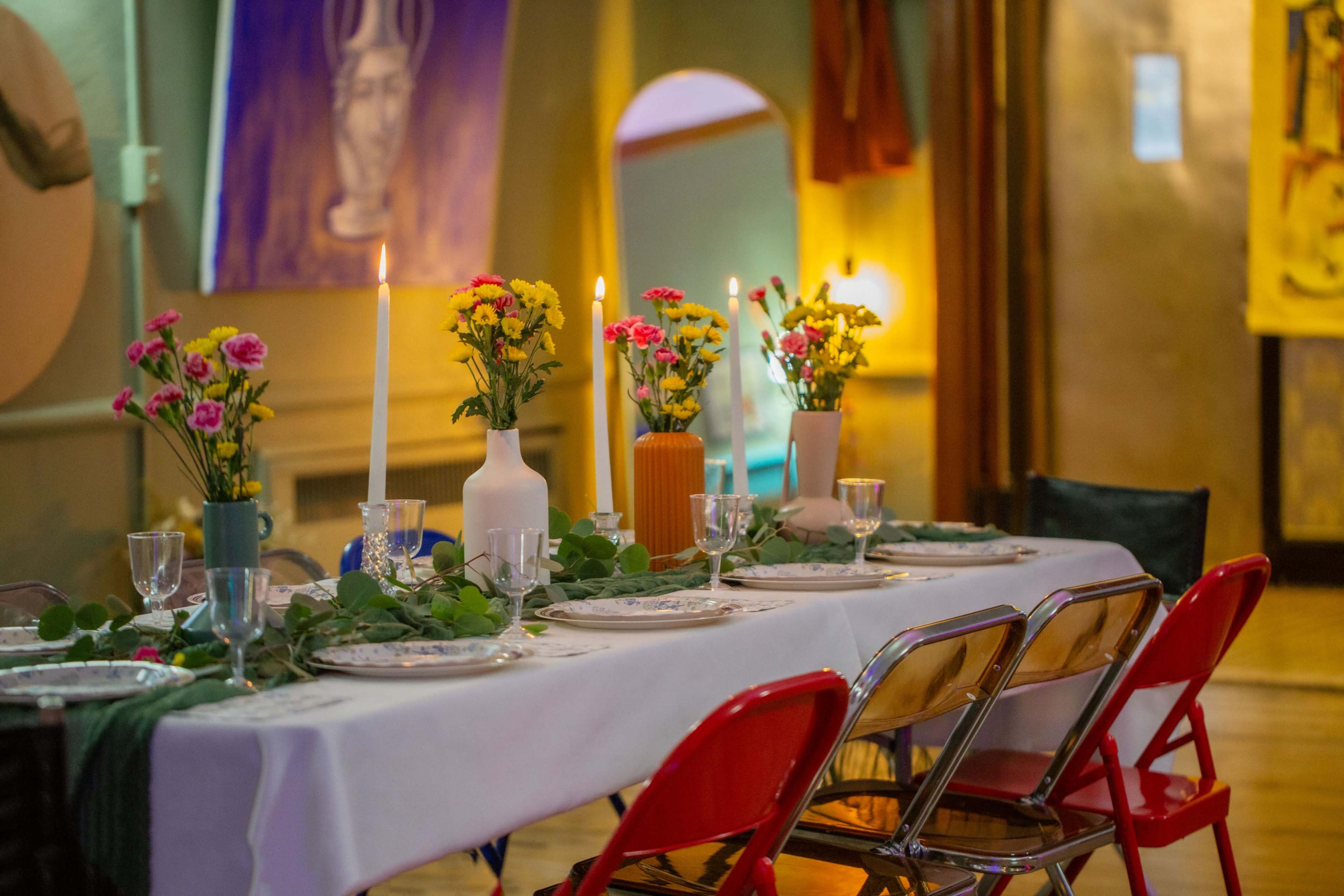 A long table is set with white tablecloths, candles, floral arrangements, and glassware in a warmly lit room adorned with artwork.