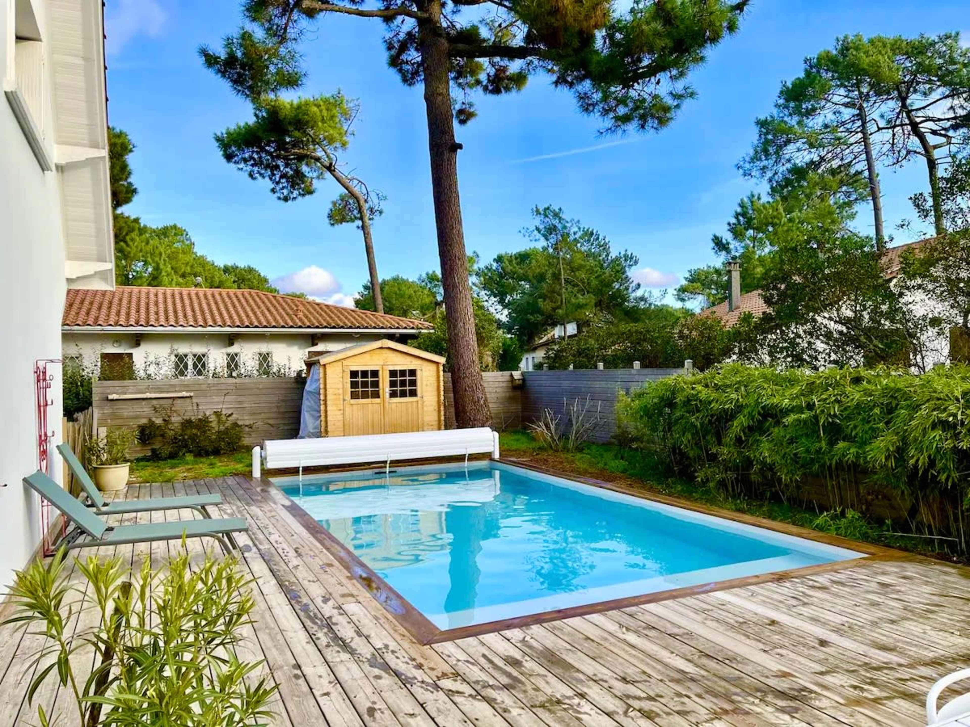The image shows a rectangular swimming pool surrounded by wooden decking, with a small wooden shed and tall trees in the background.