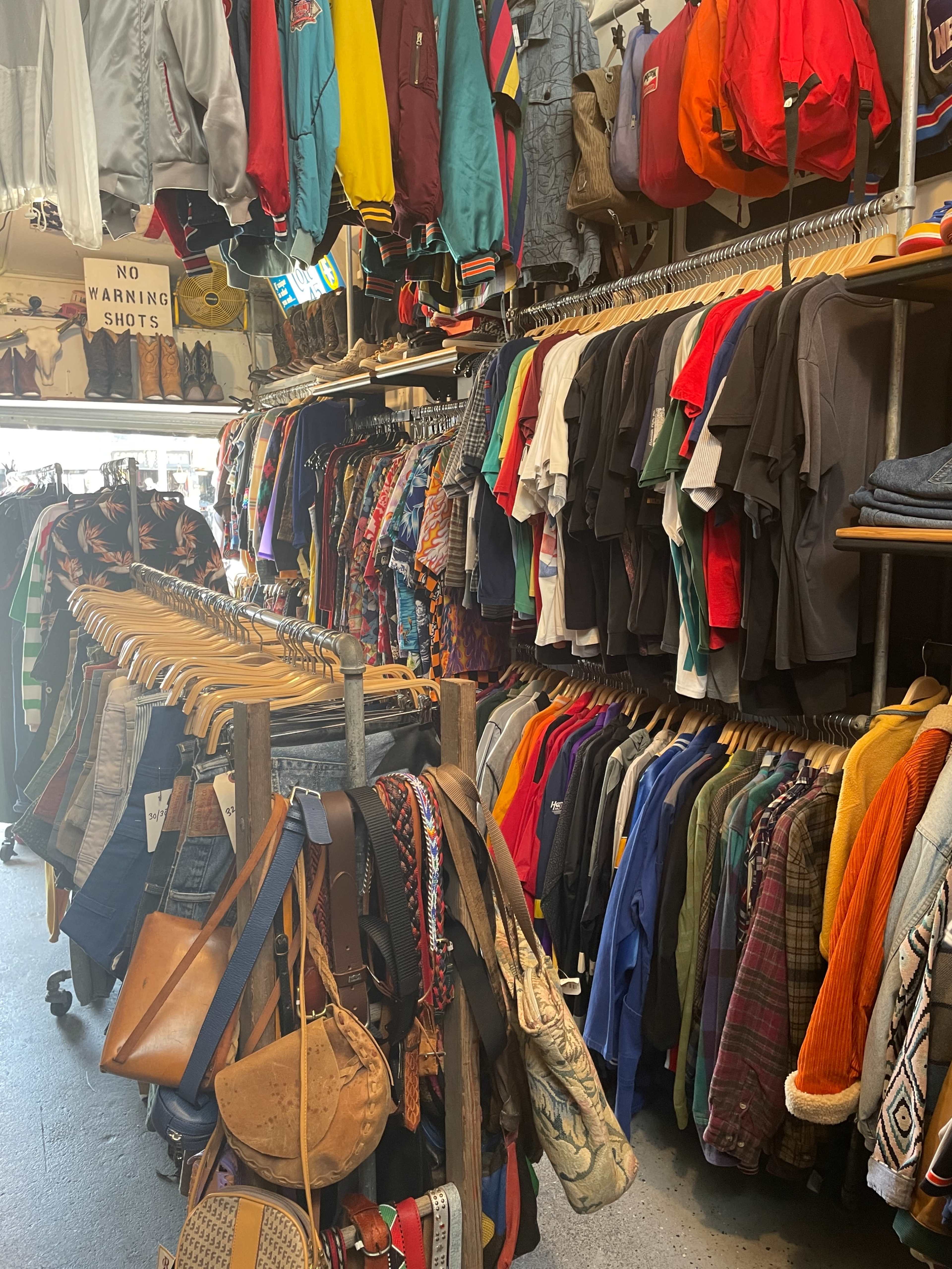 The image shows a cluttered thrift store aisle filled with hanging clothing items and accessories organized on racks.