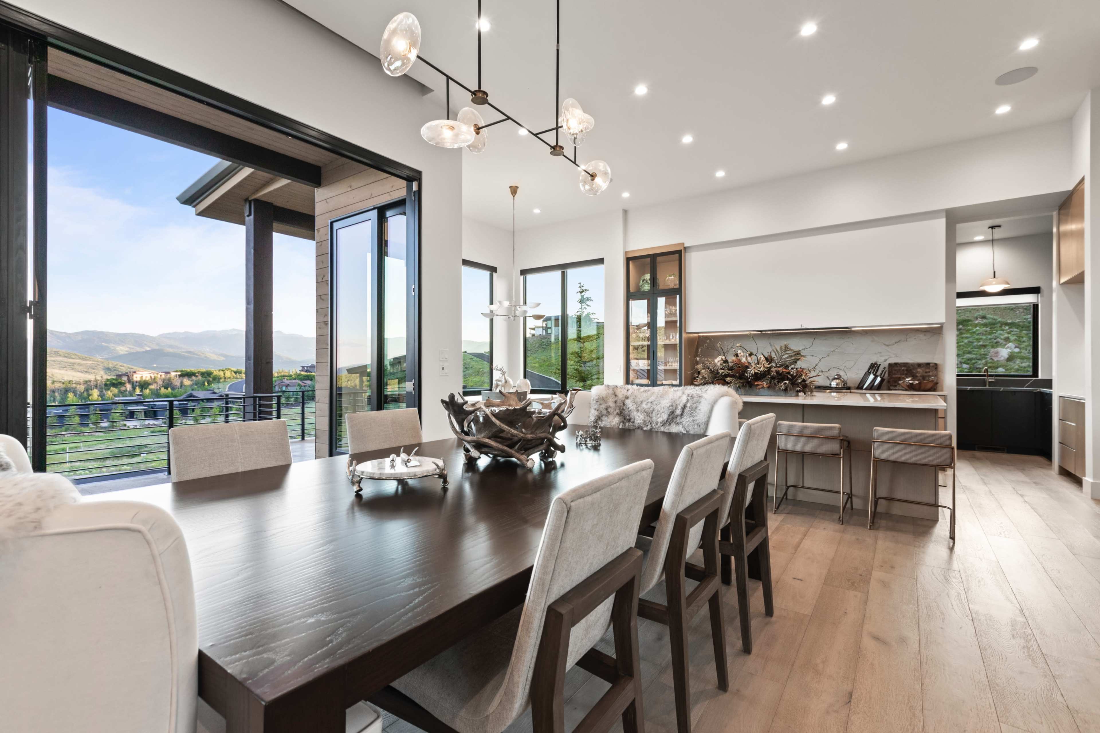 A modern dining area with a large wooden table, stylish chairs, and large windows offering a view of the landscape outside.