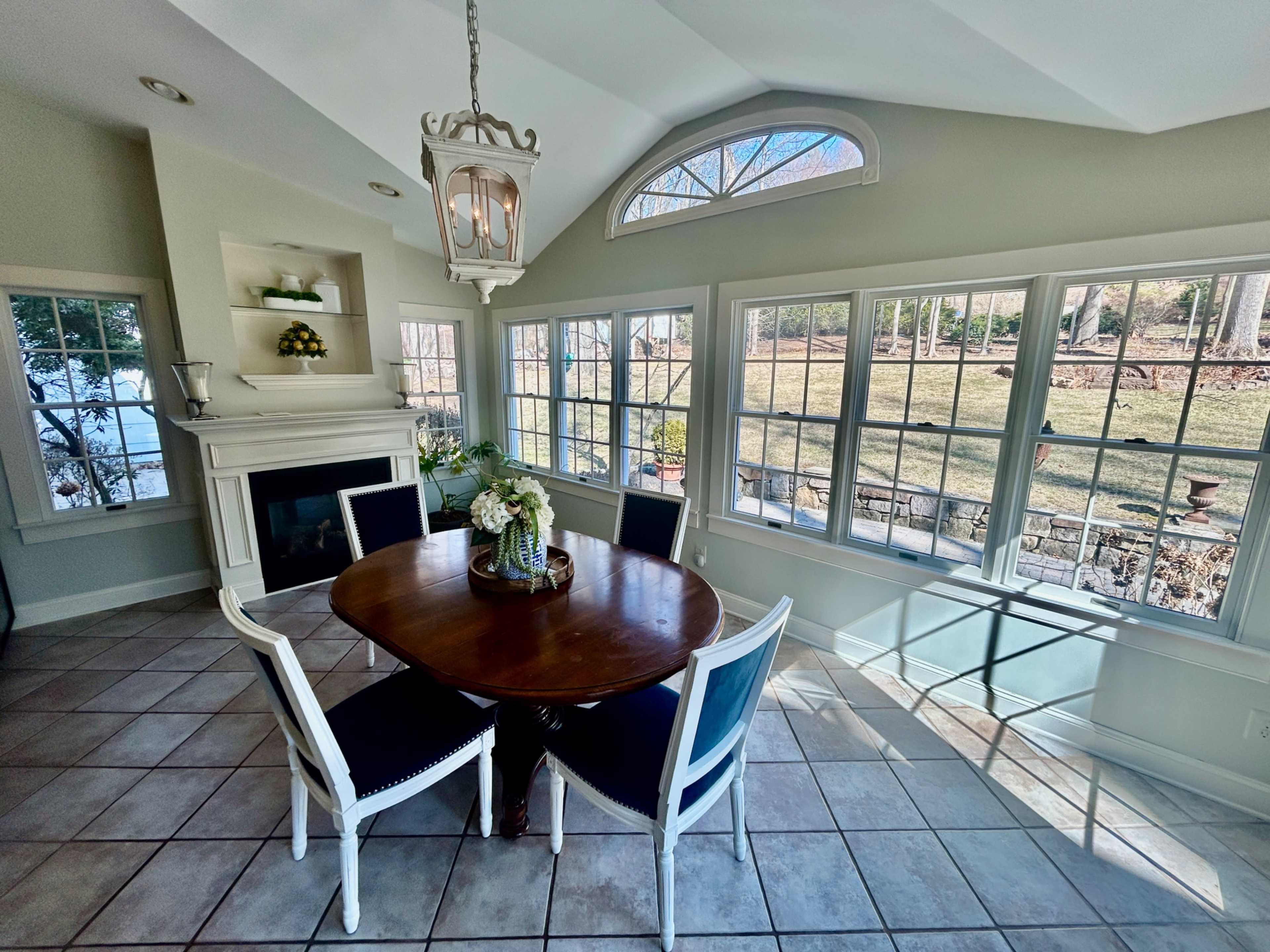 A dining area features a round wooden table surrounded by white and blue chairs, with large windows offering a view of the outdoors and a fireplace on one wall.