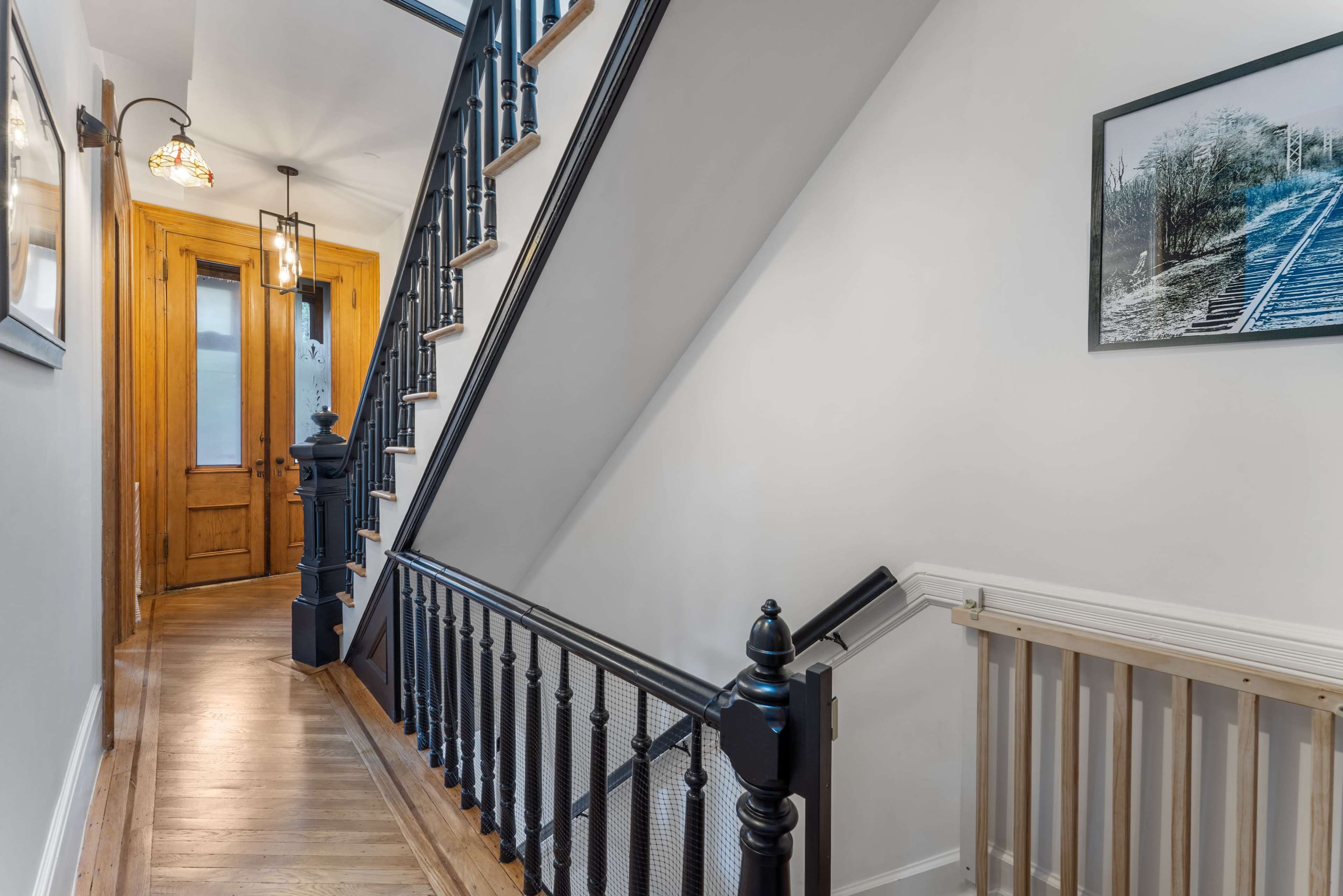 The image shows a well-lit hallway featuring a staircase with black railings, wooden steps, and a door at the end, complemented by artwork on the walls.