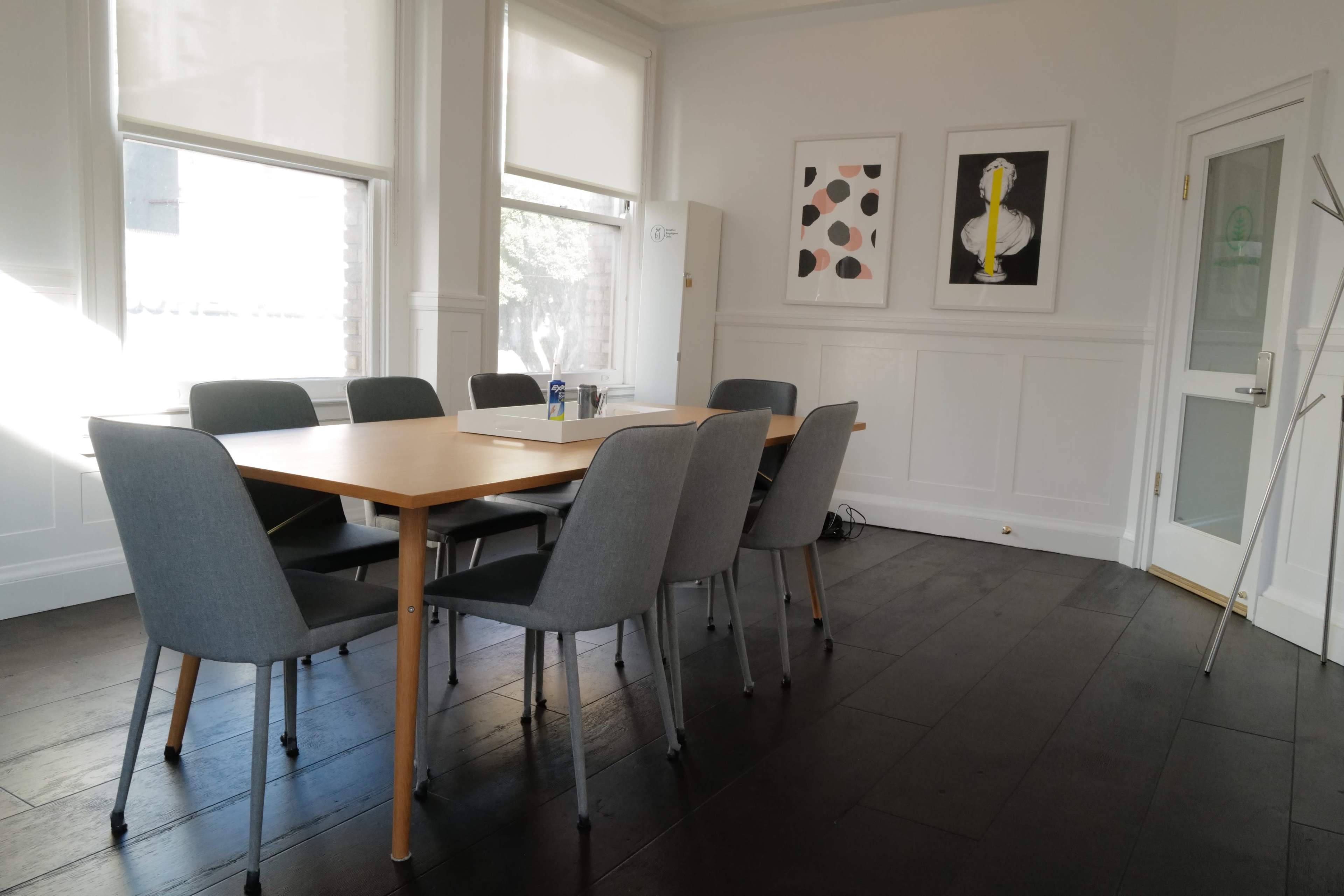 A wooden conference table surrounded by gray chairs is set in a brightly lit meeting room with large windows and two framed artworks on the walls.