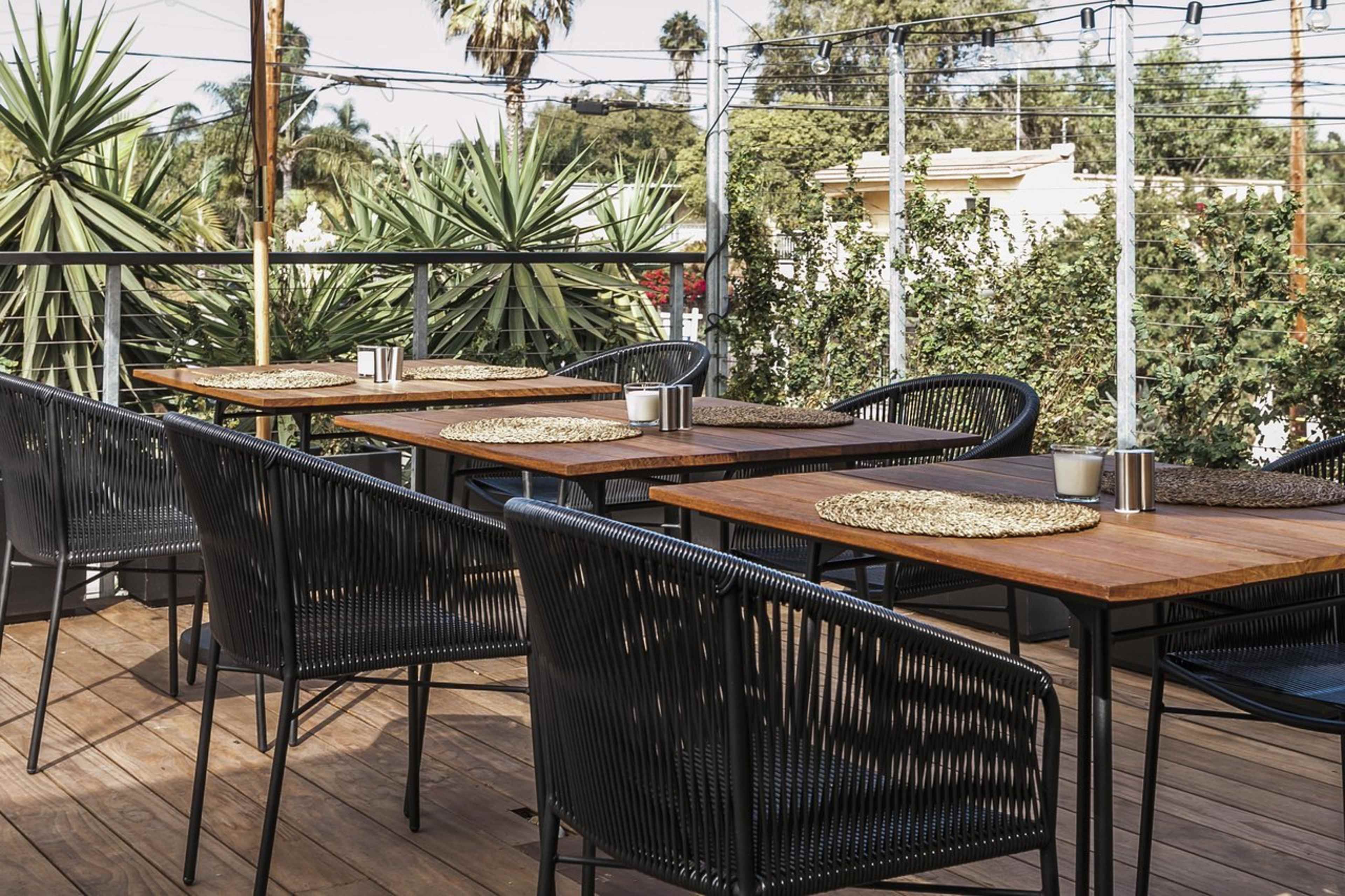 A set of tables and chairs arranged on a wooden deck, surrounded by potted plants and palm trees, with power lines visible in the background.