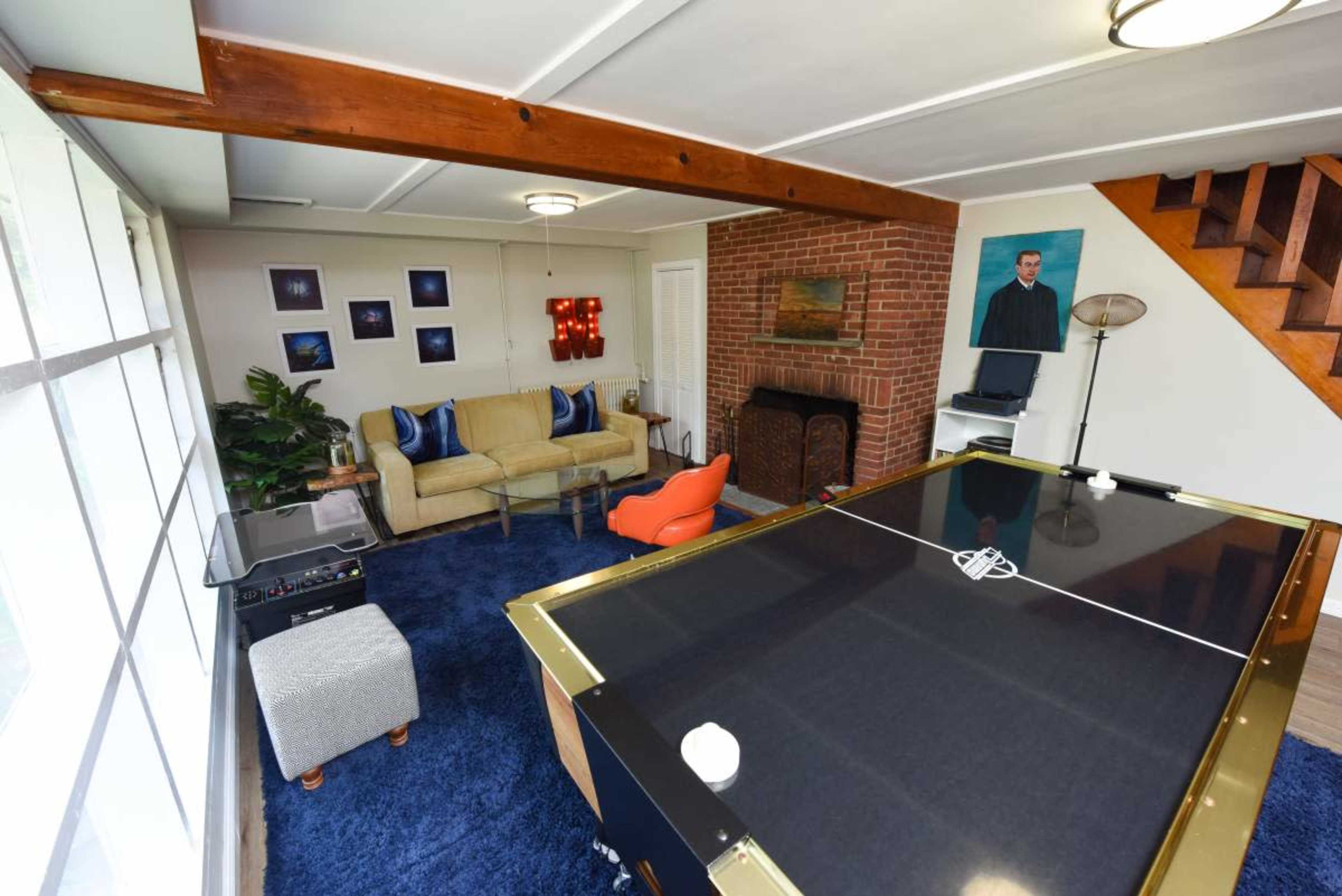 A cozy living area featuring a large air hockey table, a sofa, an orange chair, and decor including a brick fireplace and wall art.