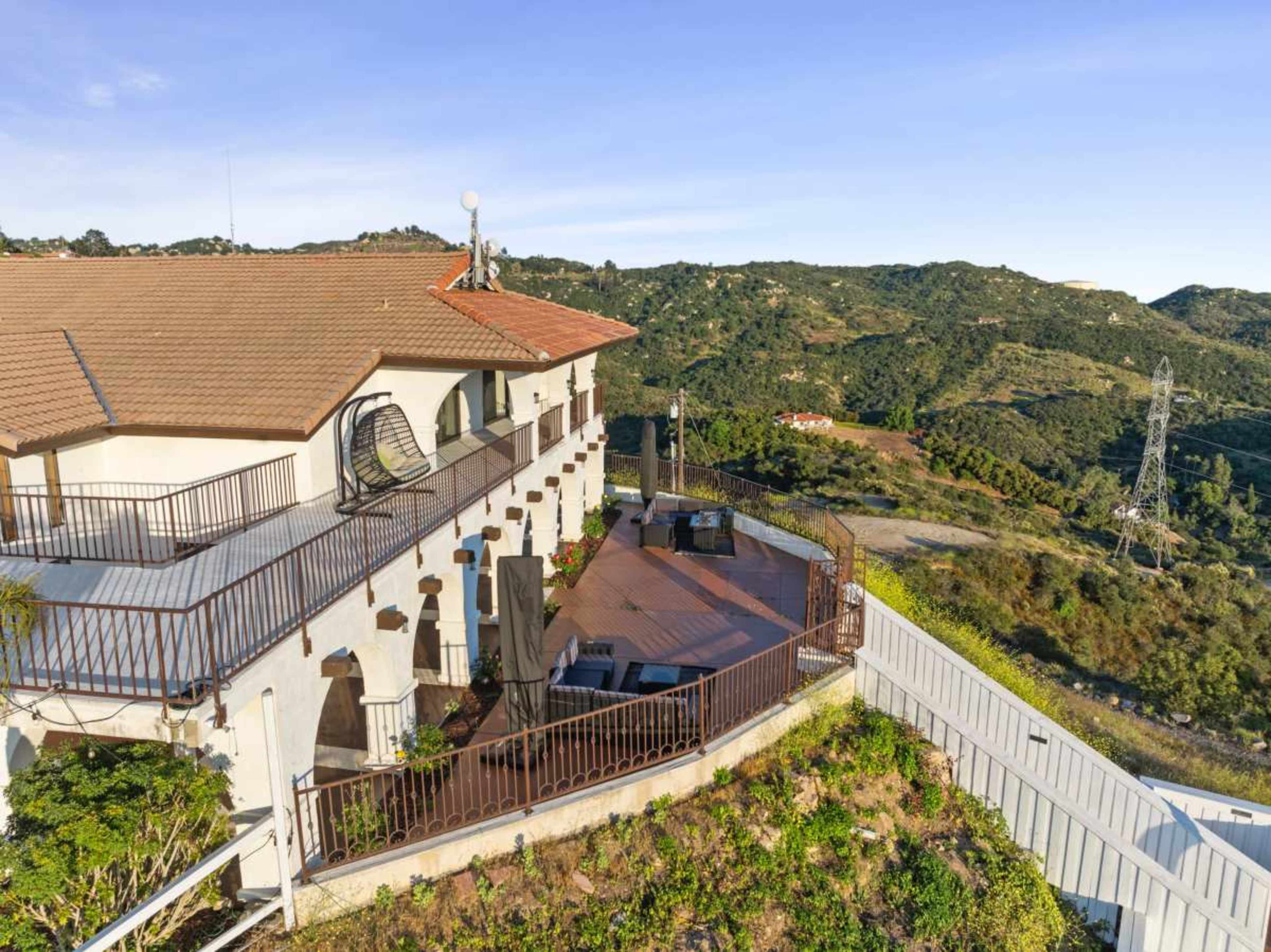 A large hillside home features a spacious deck overlooking a rolling green landscape with distant hills and communication towers.