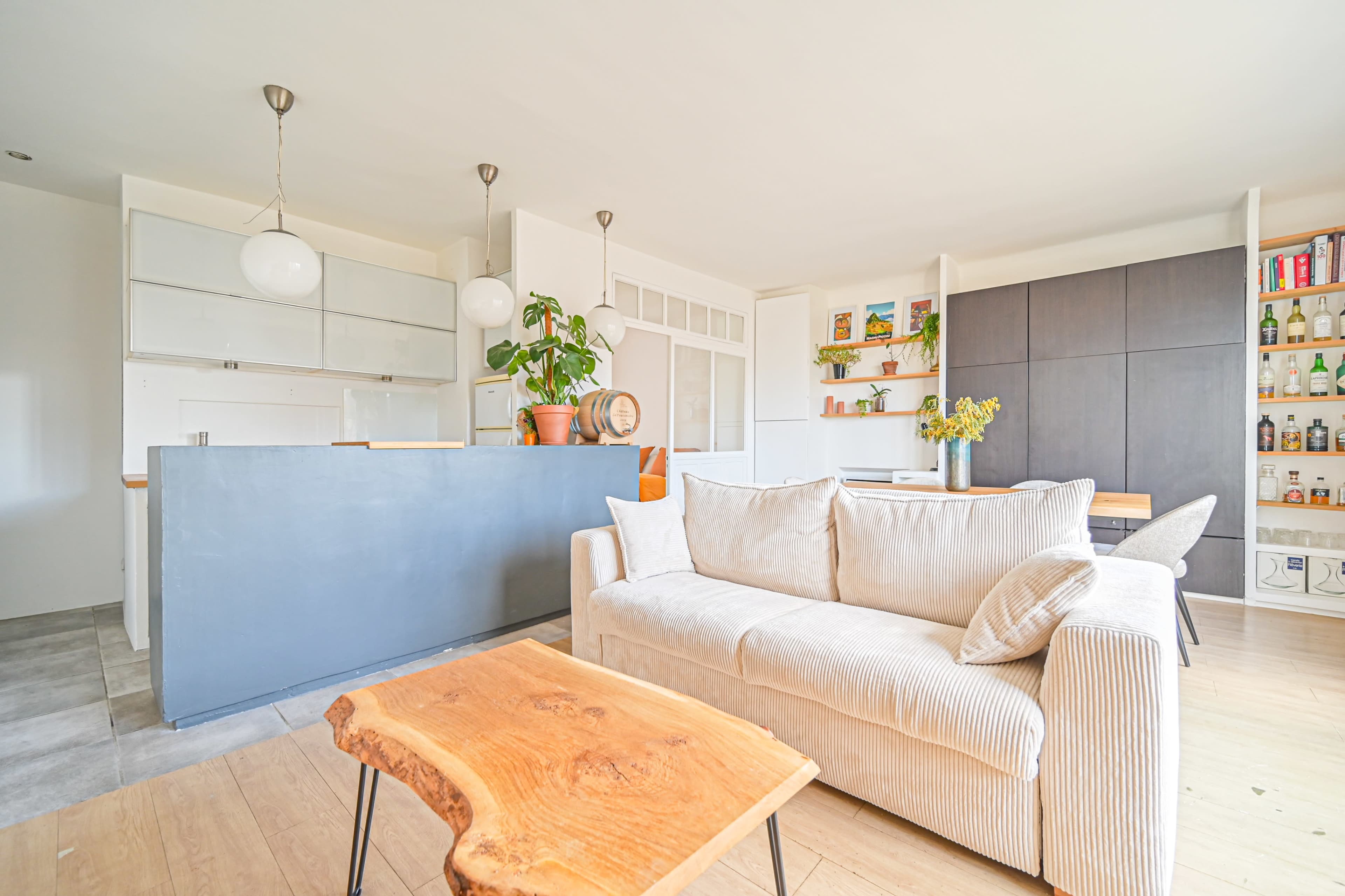 The image shows a modern living space with a light-colored sofa, a wooden coffee table, and a kitchen area featuring an island and shelves with plants and decor.