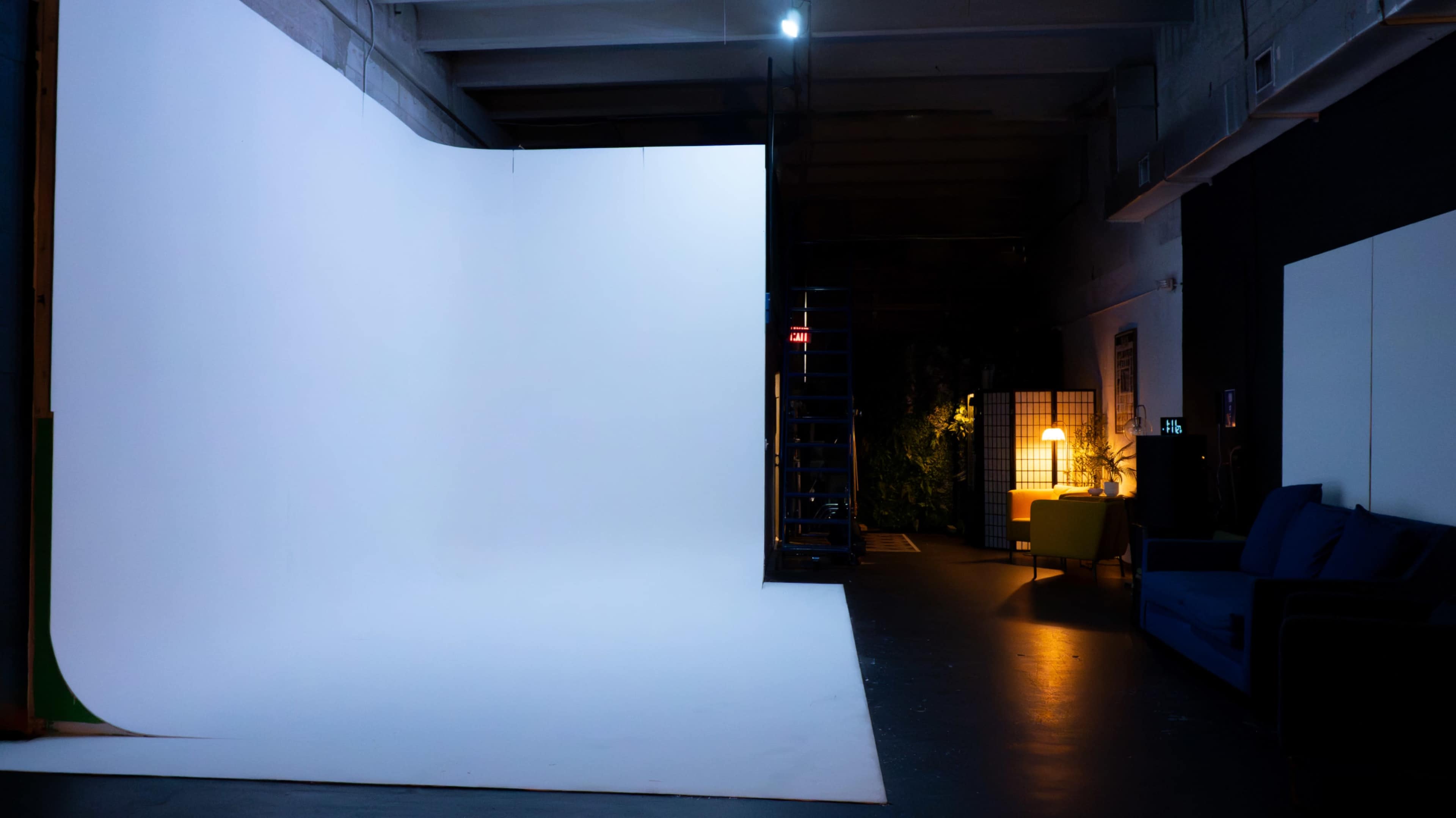 The image shows a photography studio with a large white backdrop, a ladder, and a cozy seating area featuring a lamp and a blue couch.
