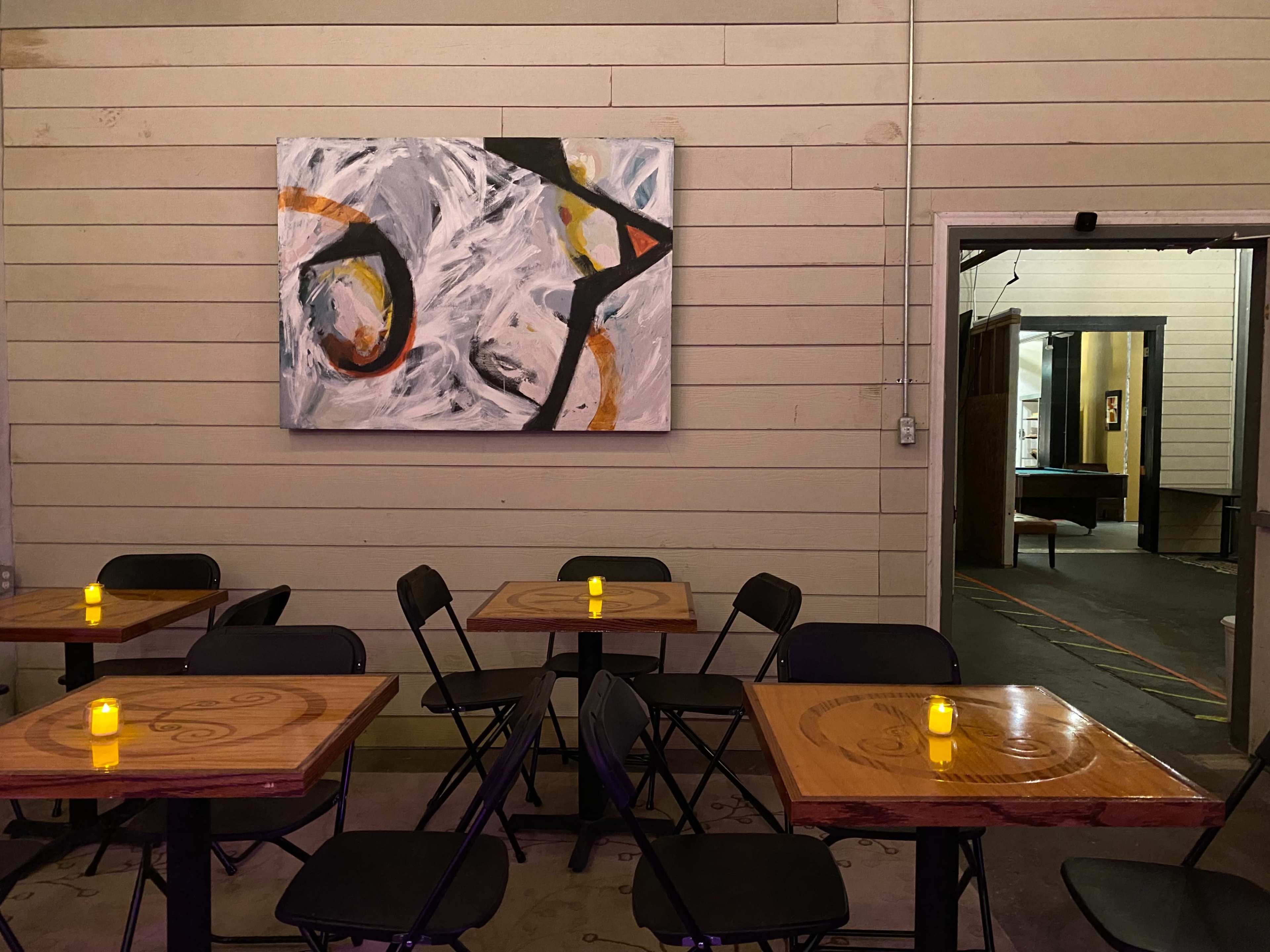 A dimly lit restaurant interior with wooden tables, black chairs, and a large abstract painting on the wall.