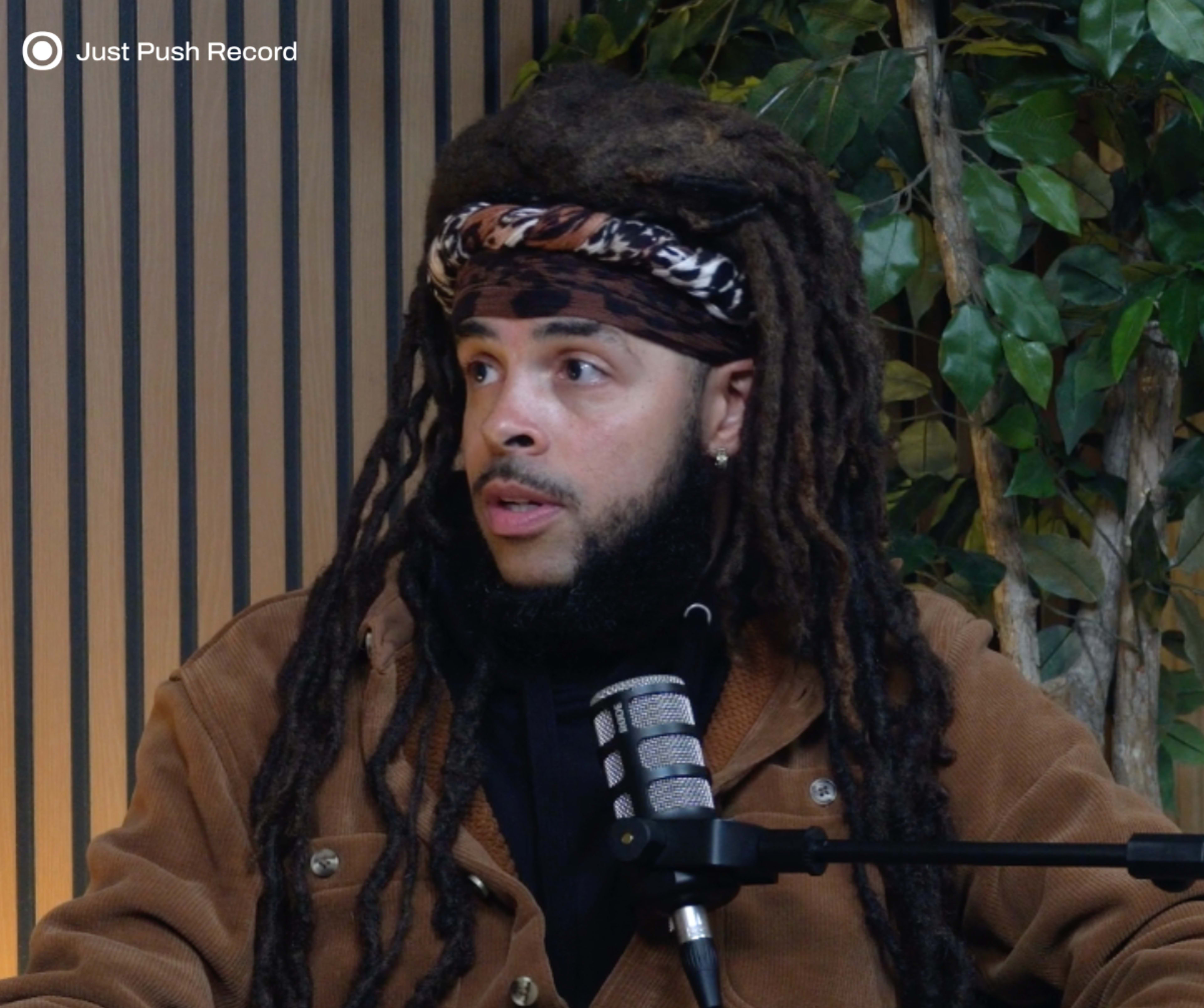 A person with long dreadlocks is sitting in front of a microphone against a backdrop of wooden panels and green plants.