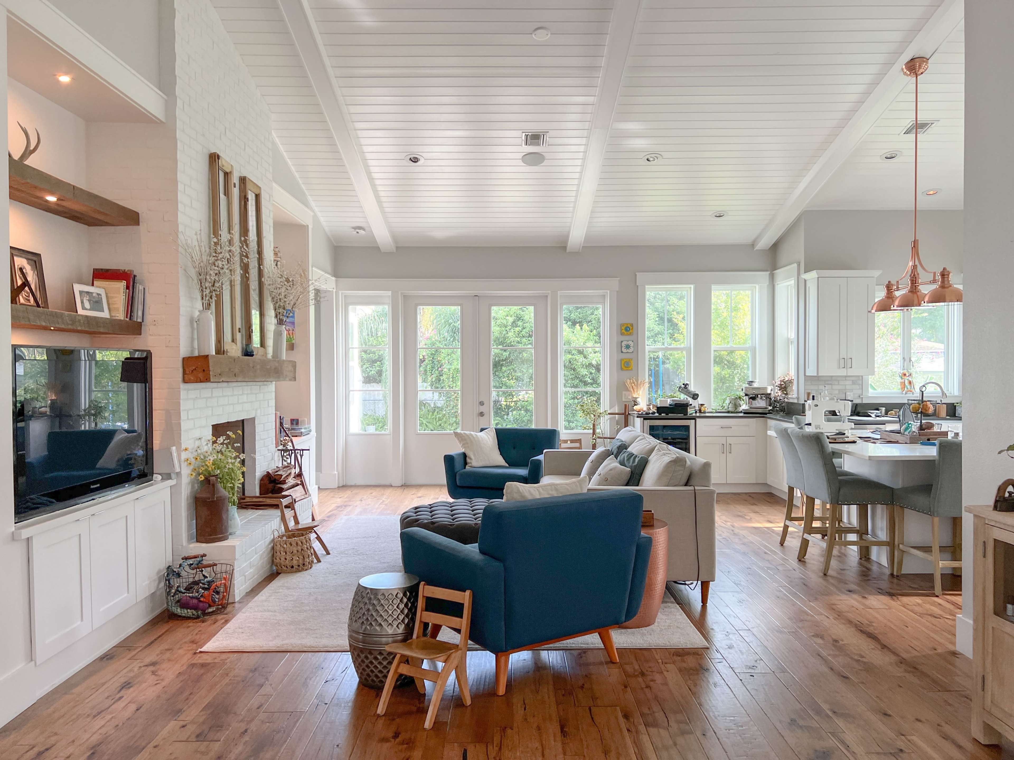 The image shows a bright, open living area with a blue and beige seating arrangement, a dining space with wooden chairs, and large windows that provide natural light.