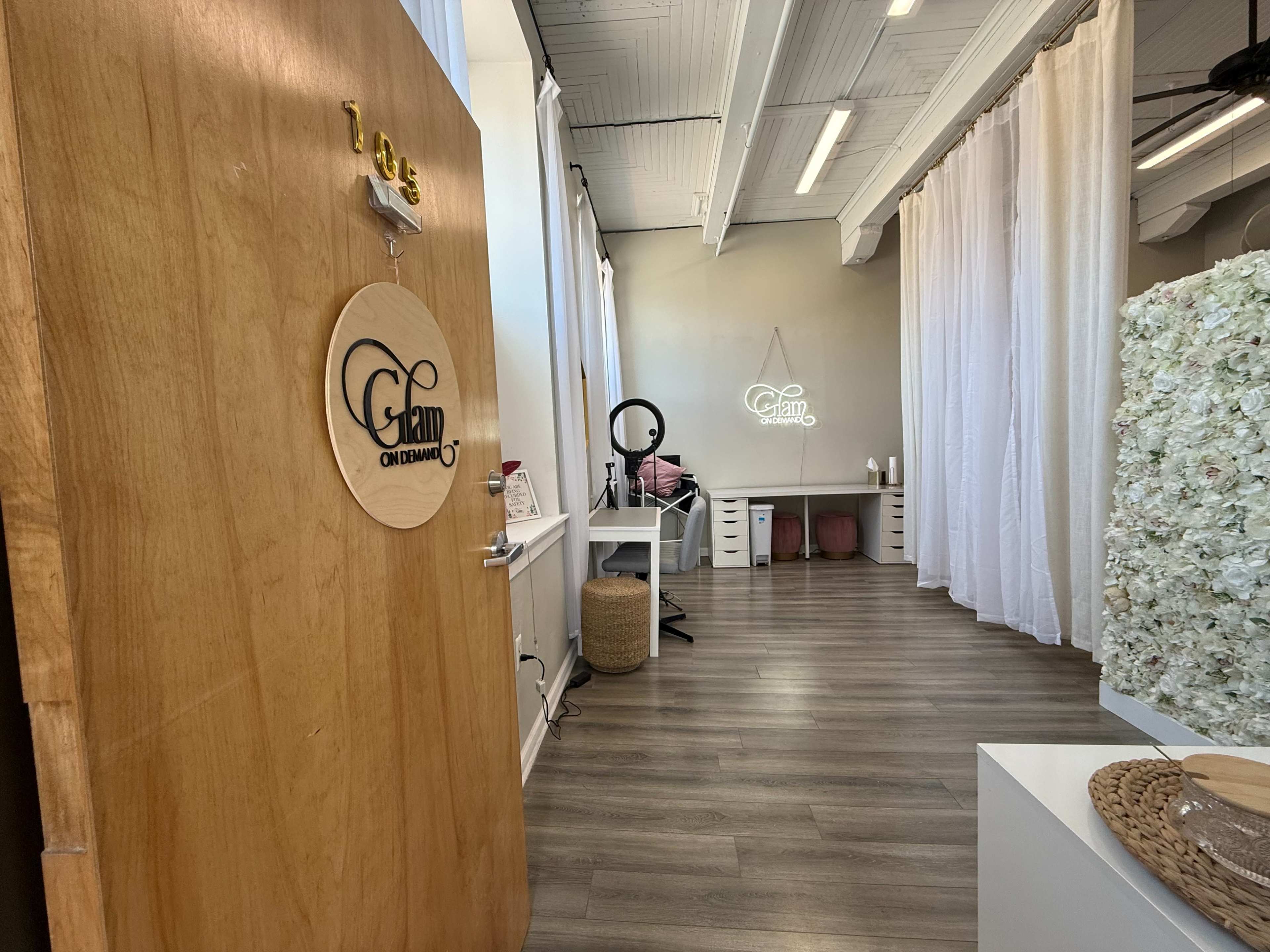 The image shows a salon or beauty studio entrance with a wooden door featuring a round sign labeled "Glam" and a clean, organized interior that includes a makeup station and decorative elements.