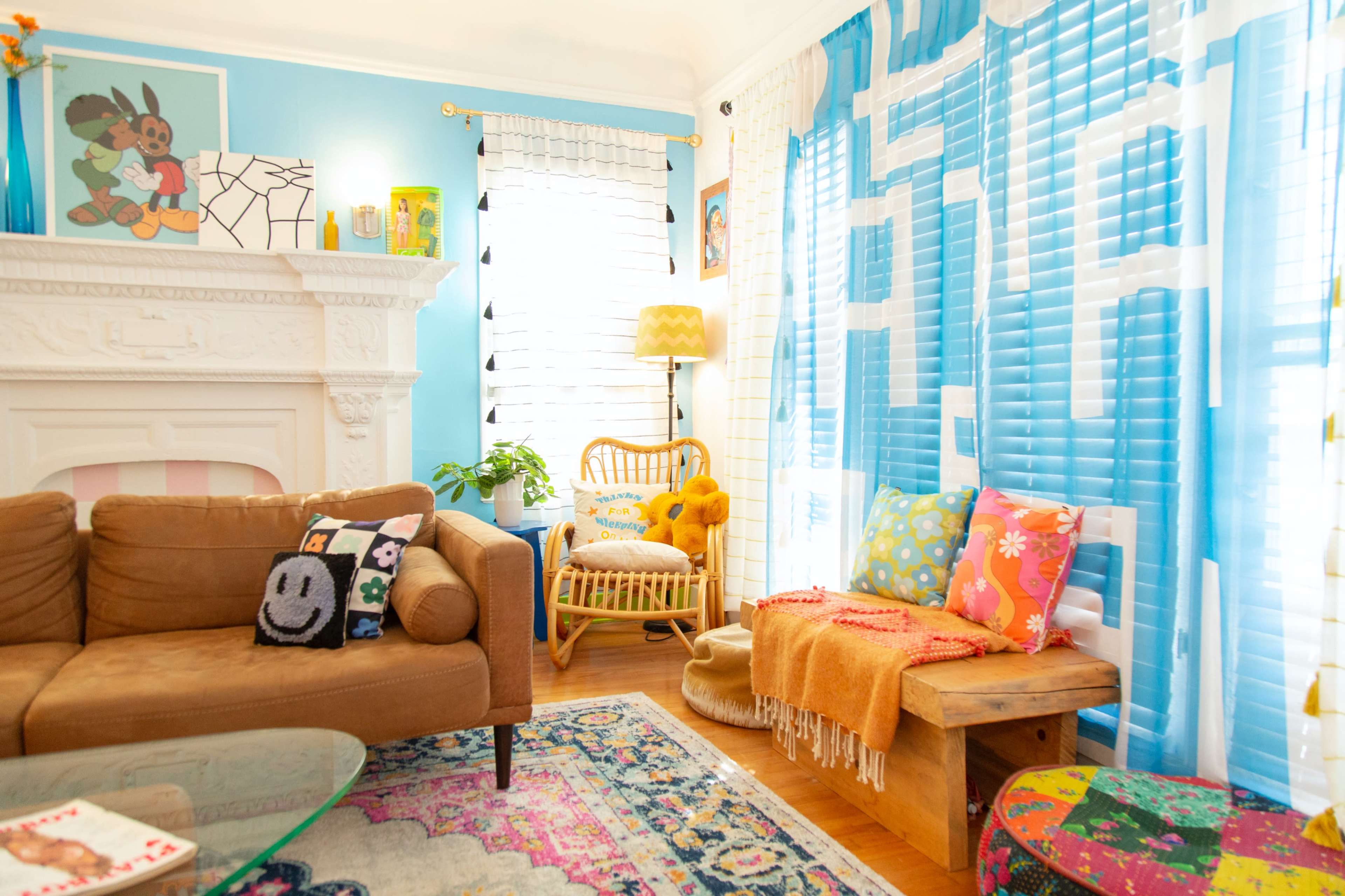 The image shows a bright and colorful living room featuring a brown sofa, a wooden coffee table, and vibrant cushions, with blue walls and large windows adorned with striped curtains.
