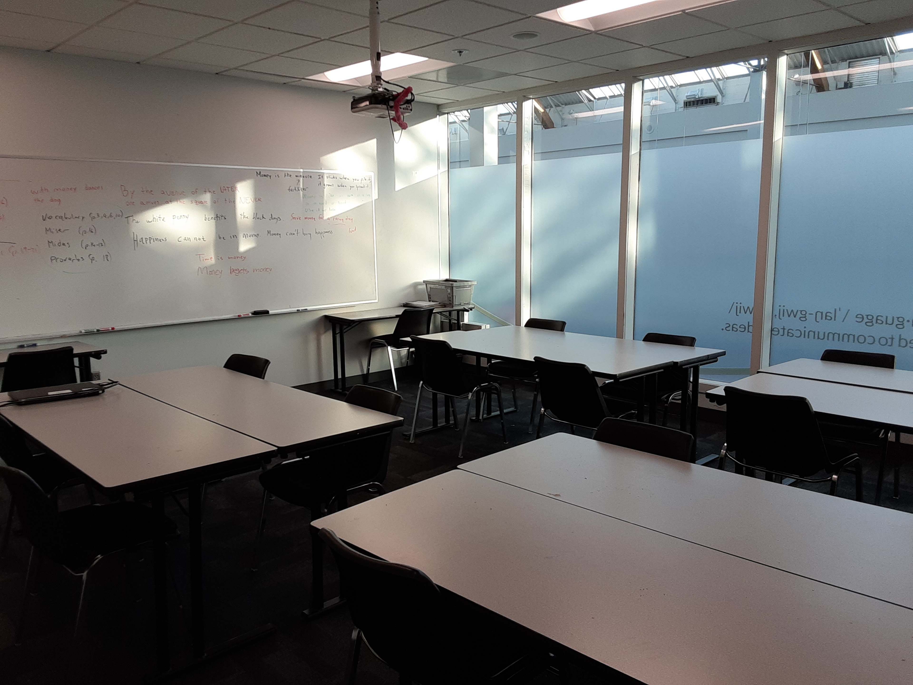 A classroom with several tables and chairs arranged around a whiteboard and large windows allowing natural light to enter.