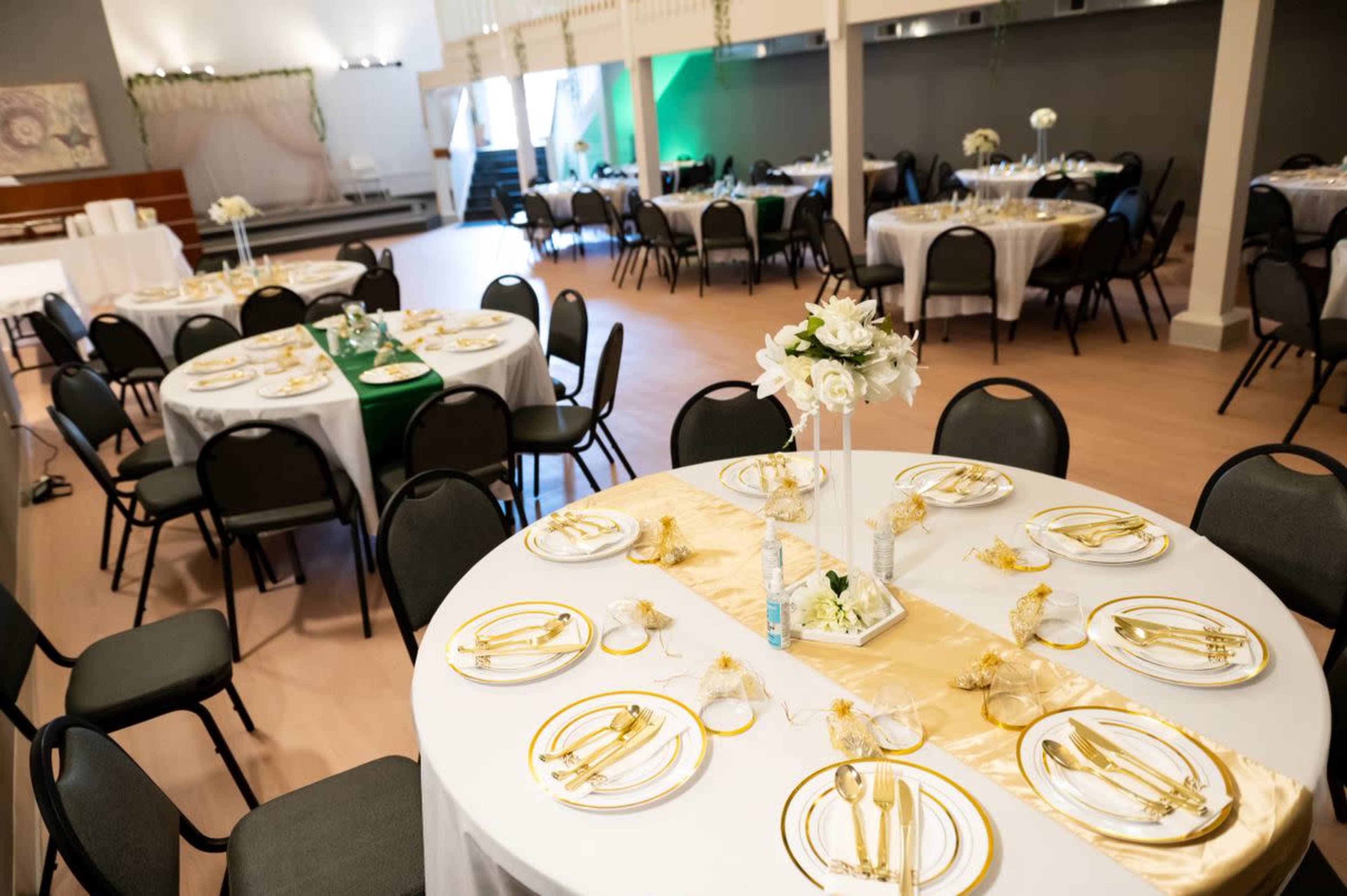 The image shows a banquet hall setup with round tables dressed in white tablecloths, each adorned with gold-trimmed dishware and floral centerpieces.