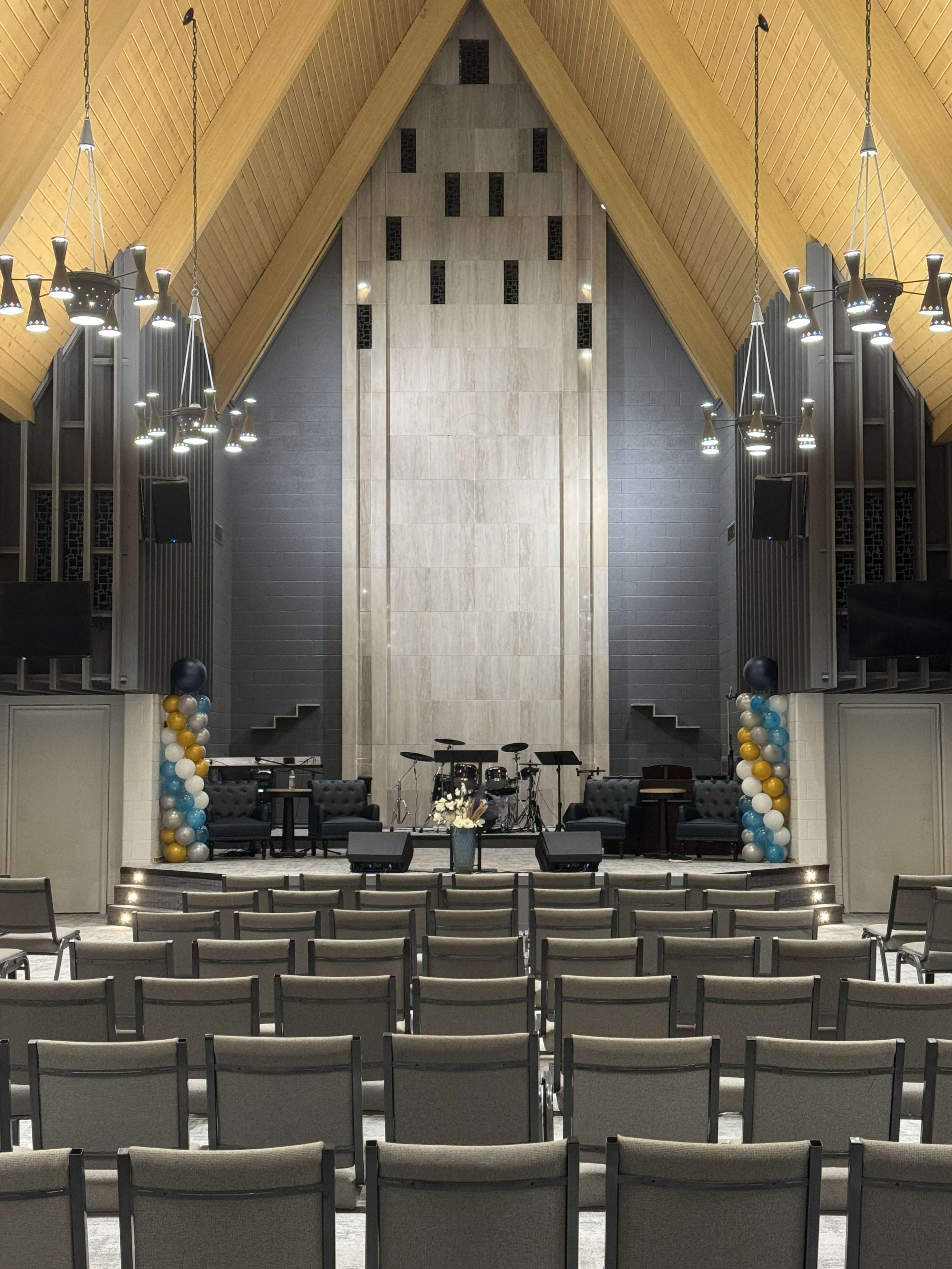 The image shows an interior view of a church sanctuary featuring rows of chairs facing a stage with musical instruments and decorative balloons at the sides.