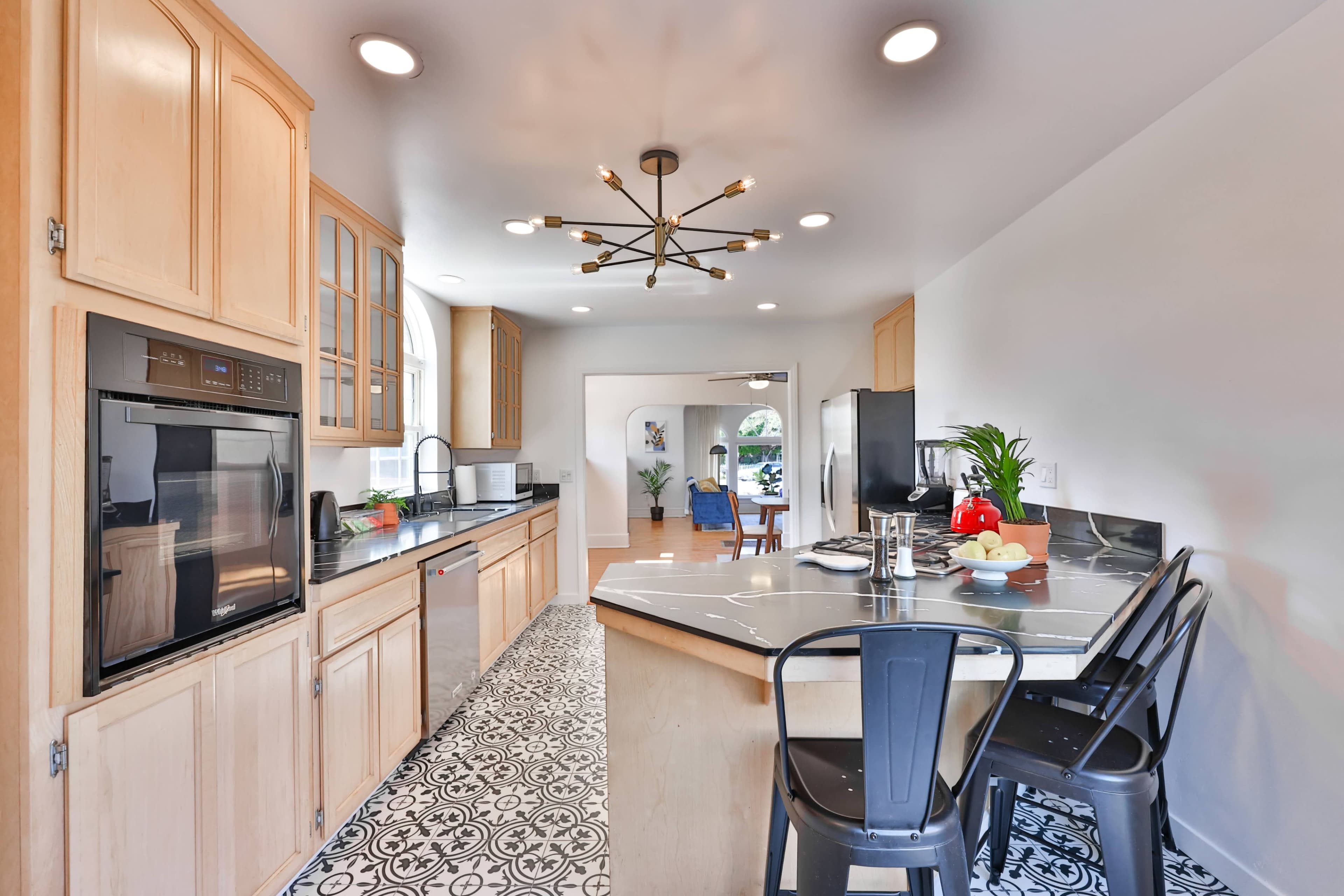A modern kitchen with wooden cabinets, stainless steel appliances, and a black and white patterned floor, featuring a countertop with two black chairs.