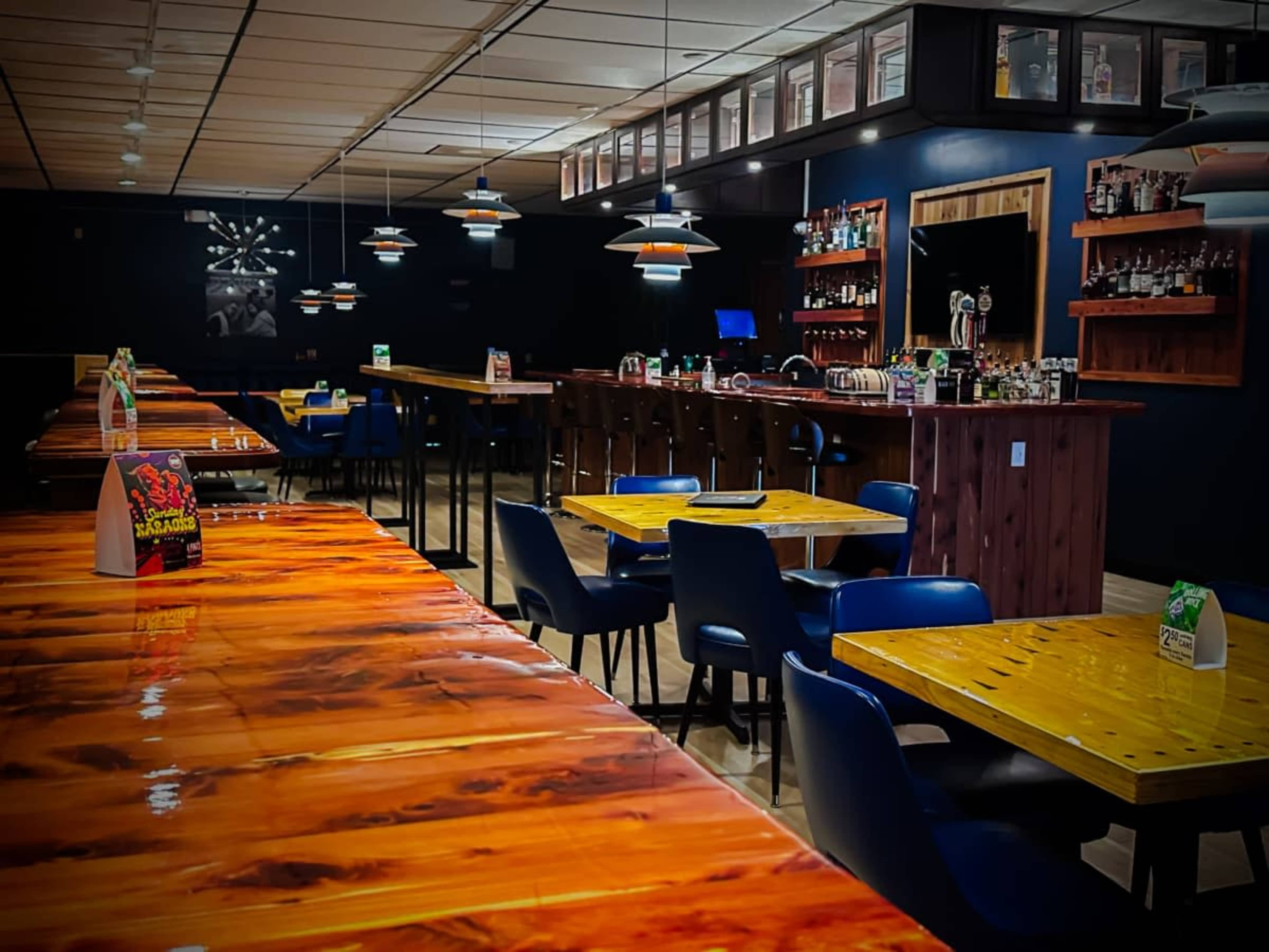 The image shows a modern restaurant interior featuring wooden tables and dark walls, with a bar area in the background illuminated by pendant lights.