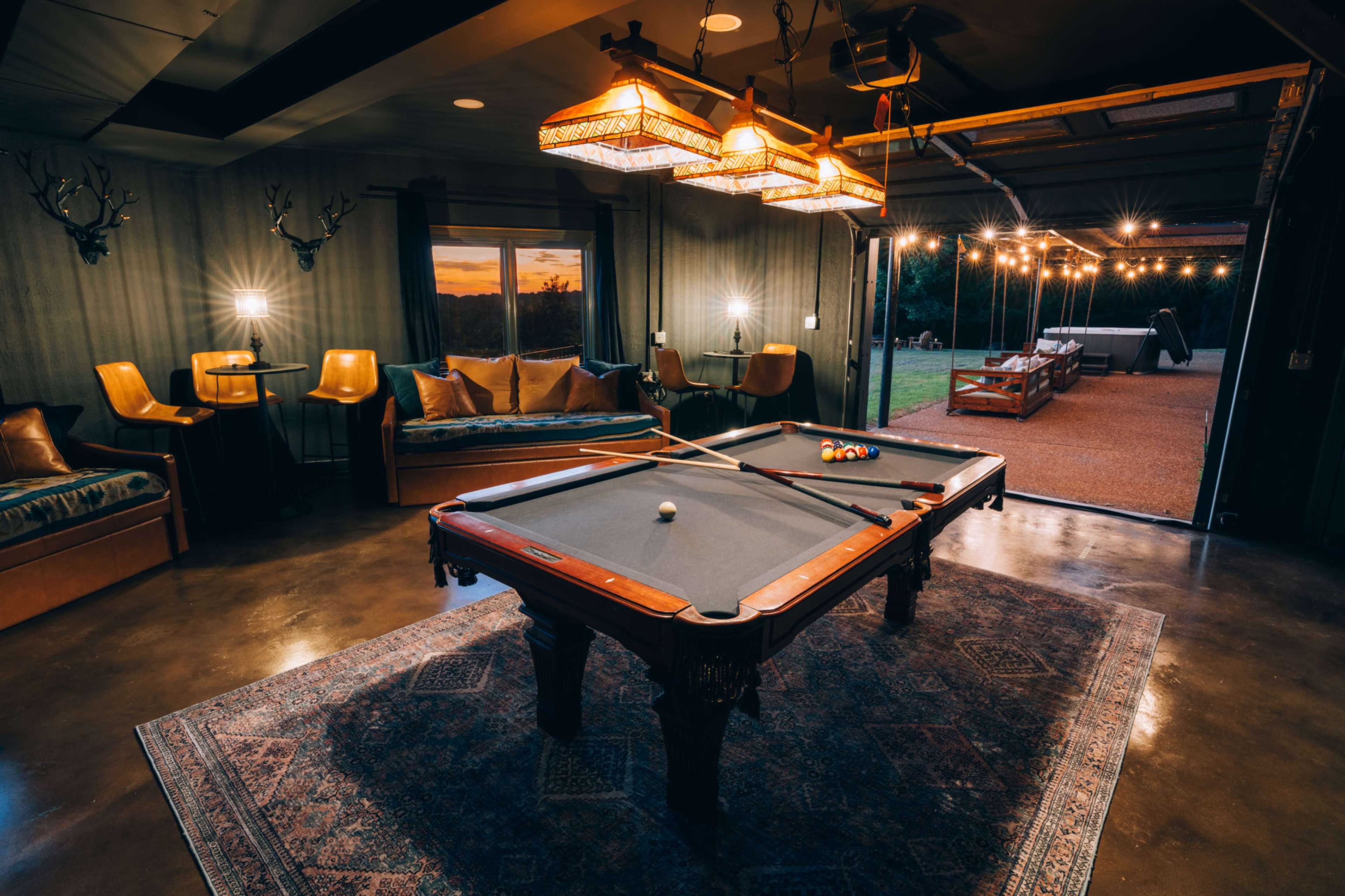 The image shows a billiards table in a dimly lit room with a view of an outdoor area illuminated by string lights.