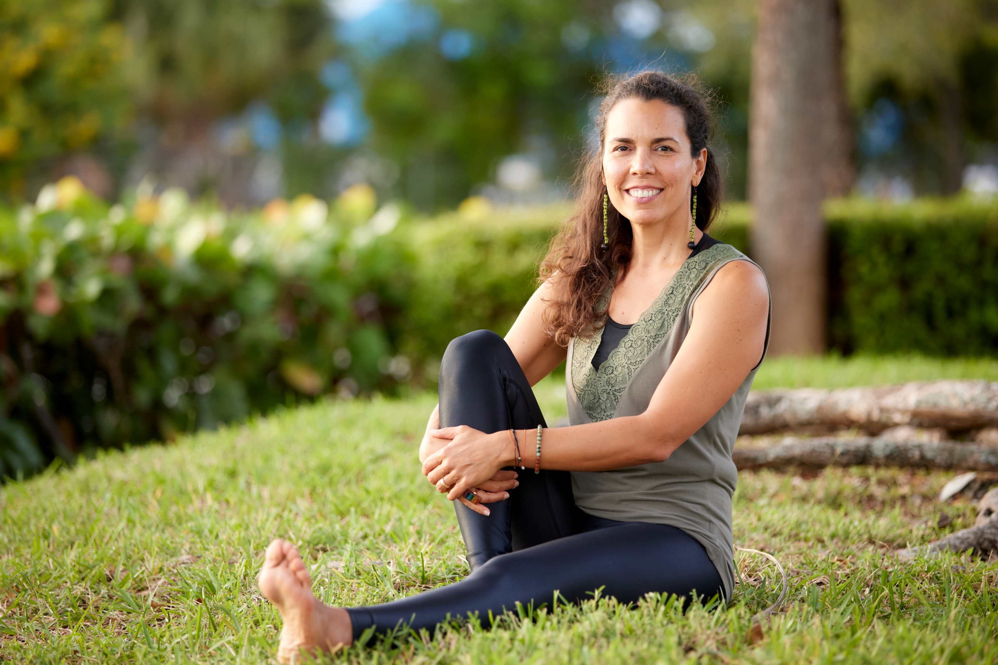A woman sits cross-legged on grass, wearing a tank top and leggings, with trees and shrubs in the background.