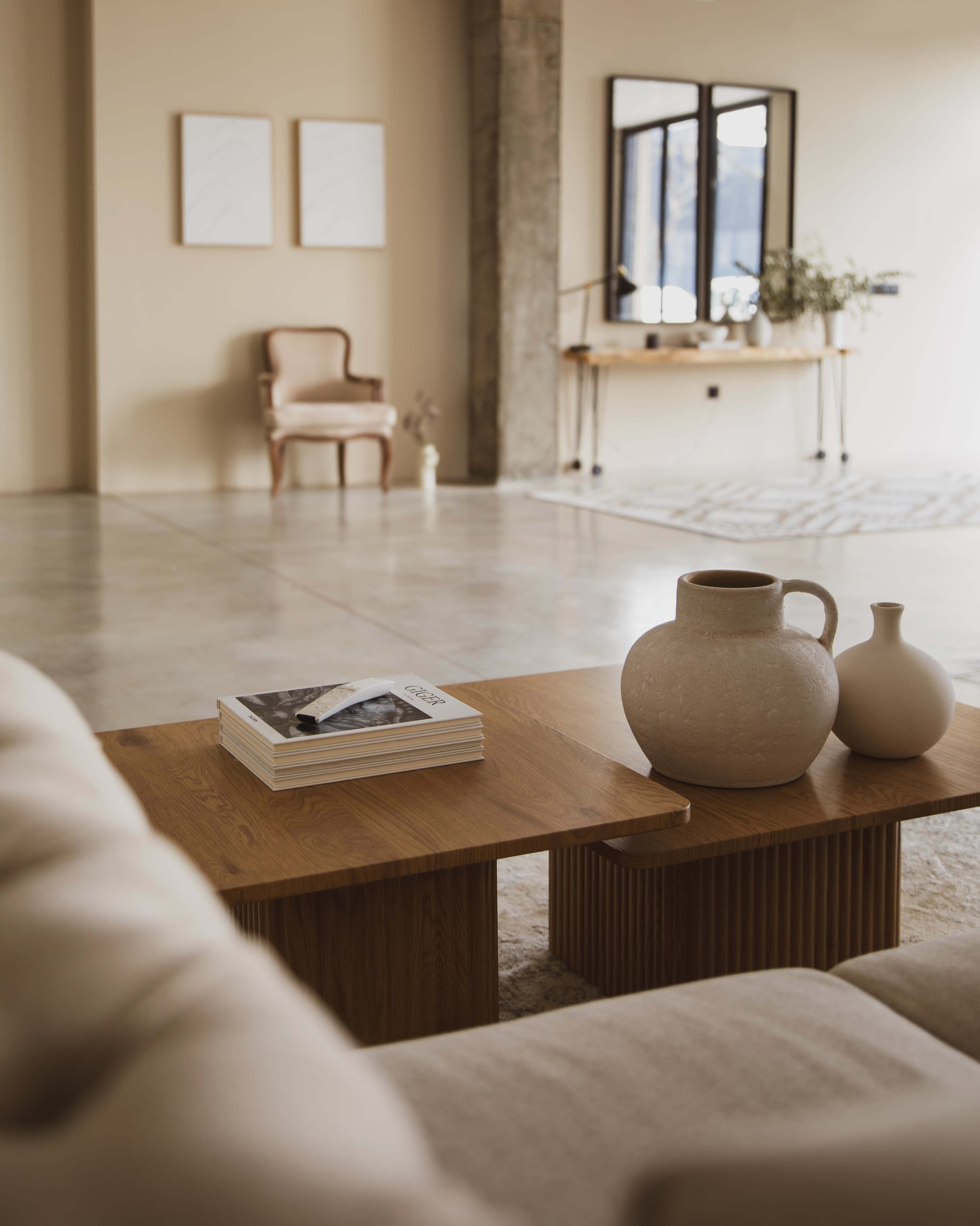 The image shows a modern interior space featuring a sofa with a coffee table topped with magazines, a ceramic pitcher, and a small vase, with a chair and a console table in the background.