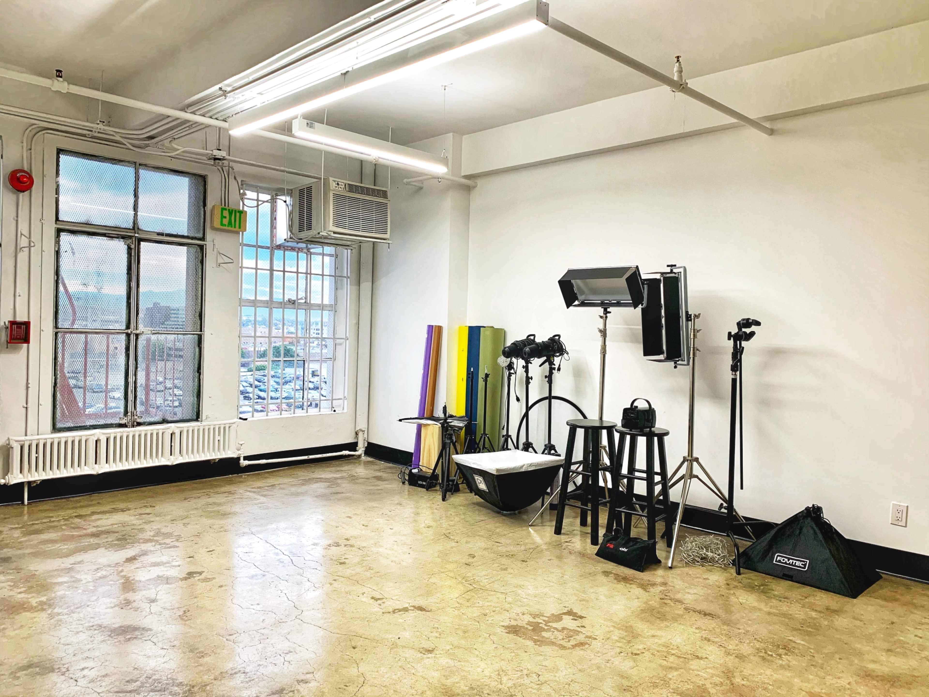A spacious, brightly lit studio with large windows, a photography setup including lights and stands, and a bare concrete floor.