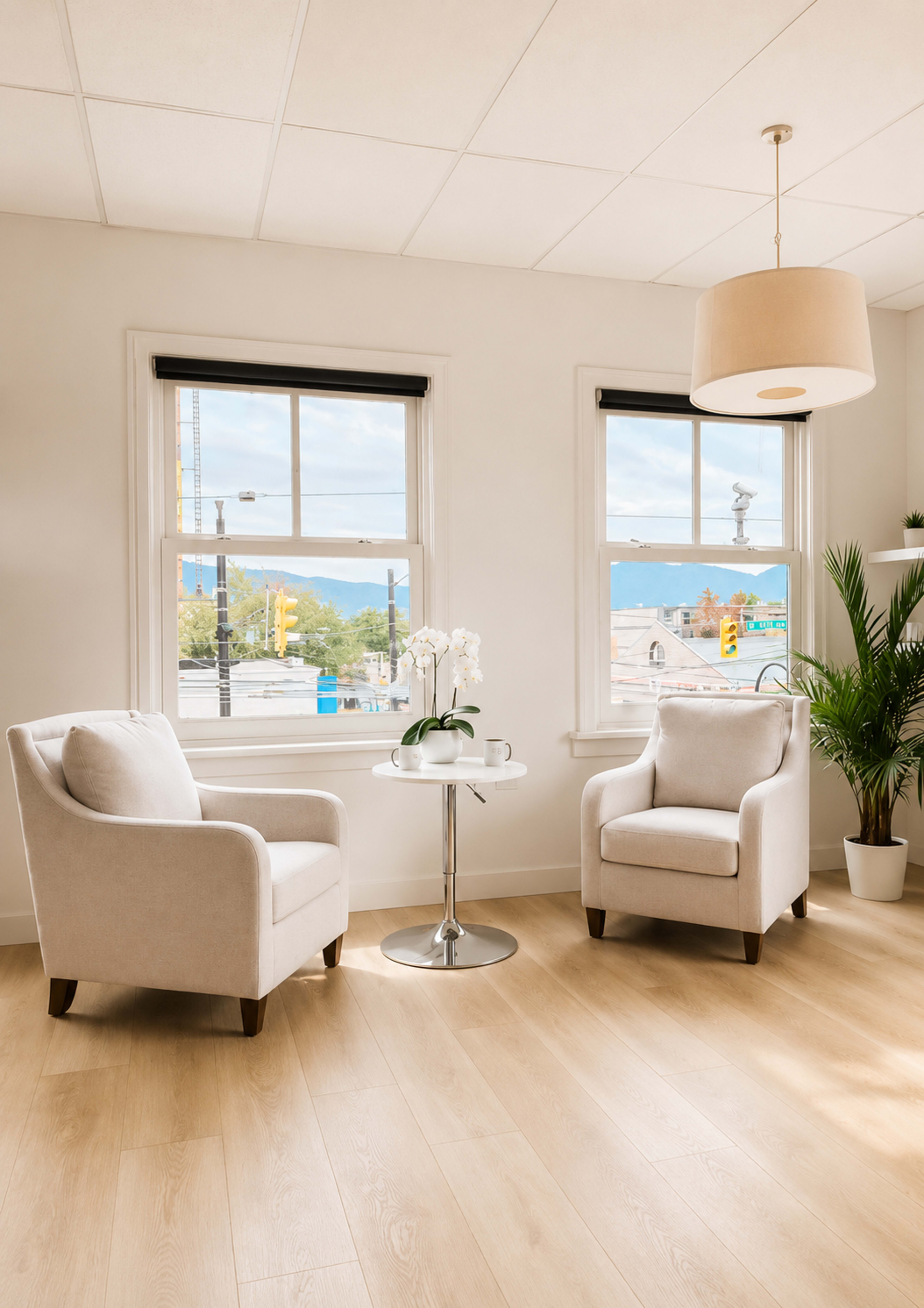 The image shows a bright room with two light-colored armchairs facing a small round table, complemented by large windows that let in natural light and offer a view of the outdoors.
