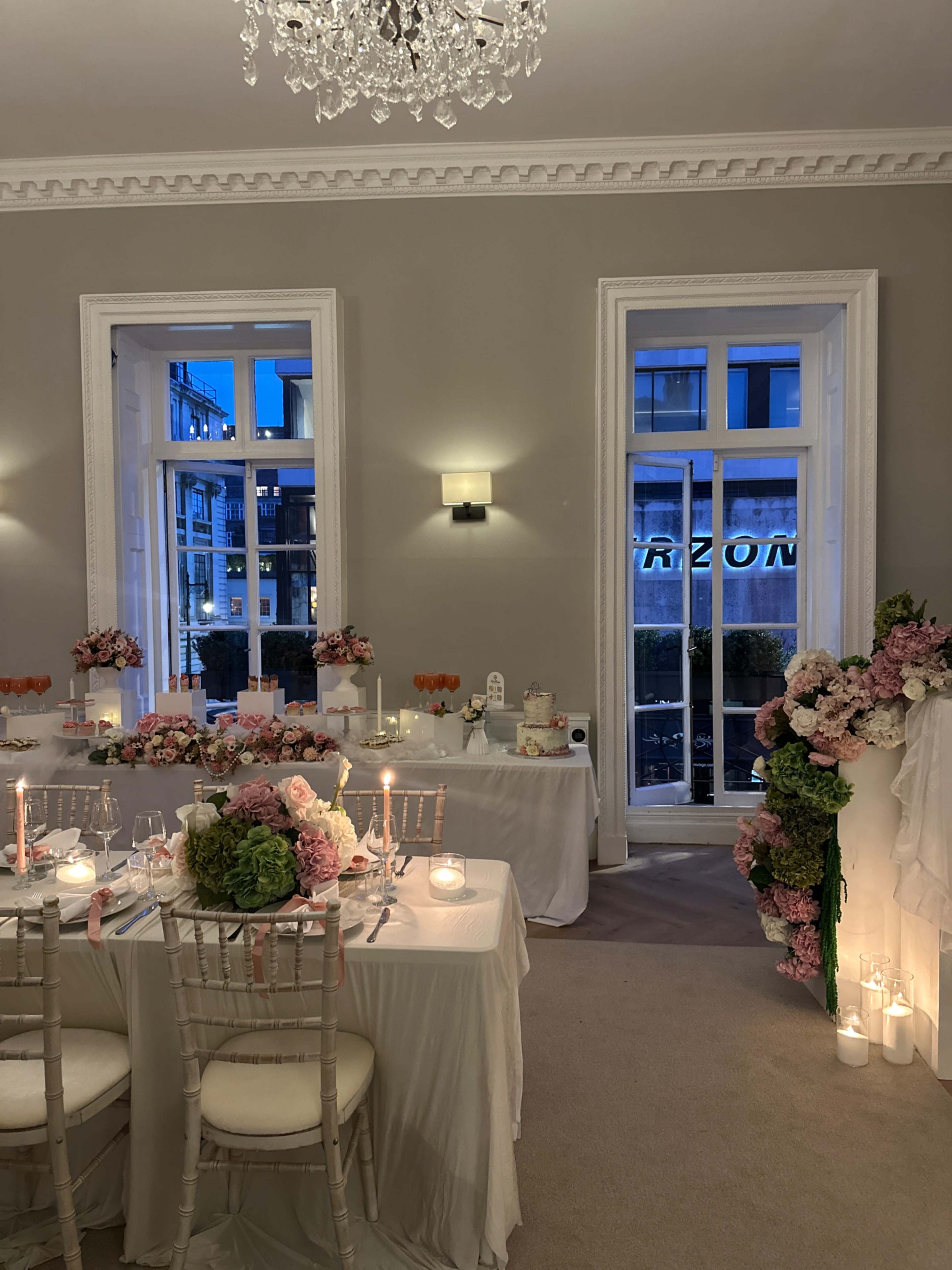 The image shows a beautifully arranged dining setup for a wedding reception, featuring elegantly dressed tables with floral centerpieces and candles, set against a backdrop of large windows.