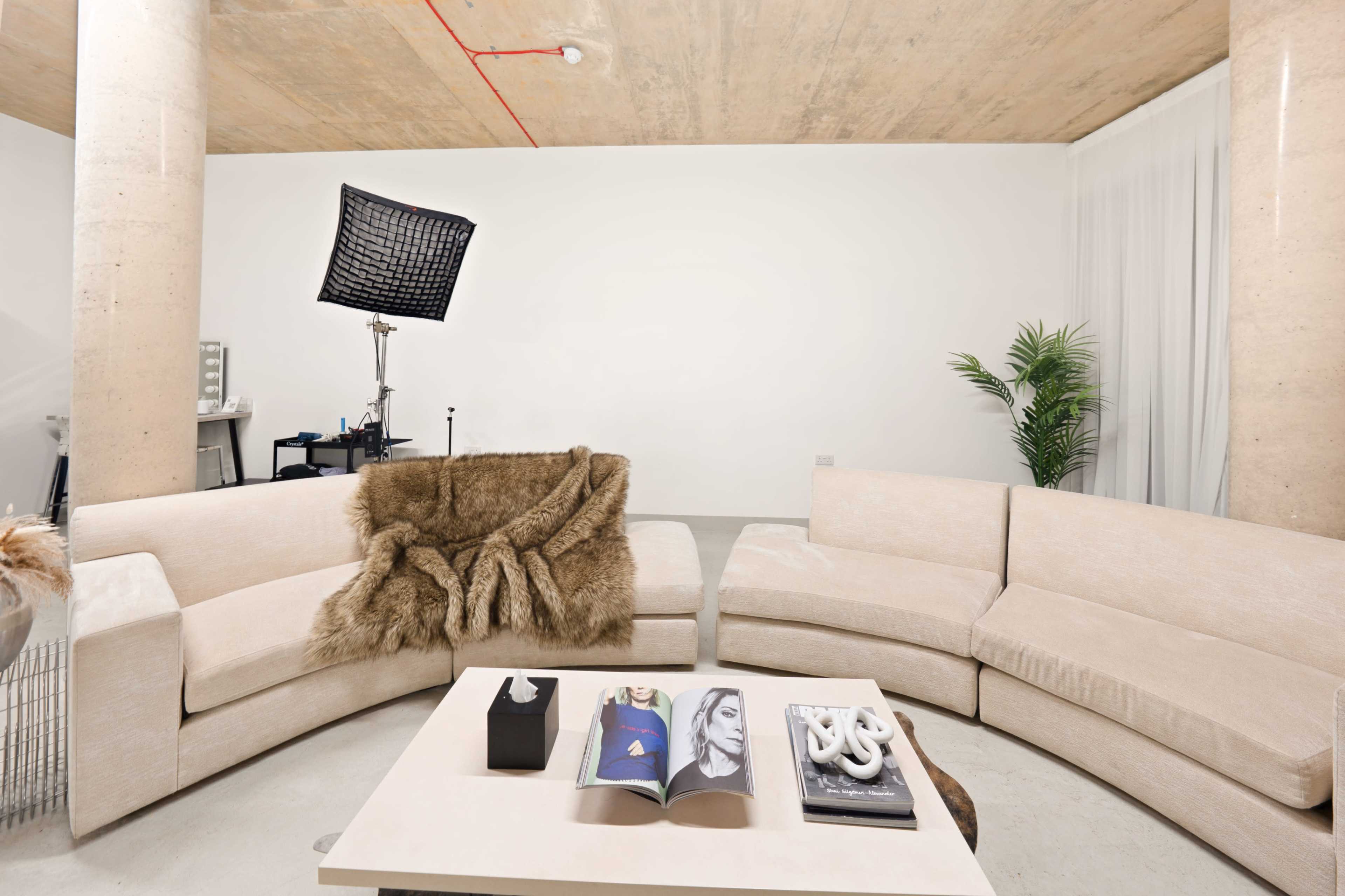 A modern living area features a curved beige sofa with a fur throw, a coffee table displaying magazines, and a backdrop stand with a light in the background.