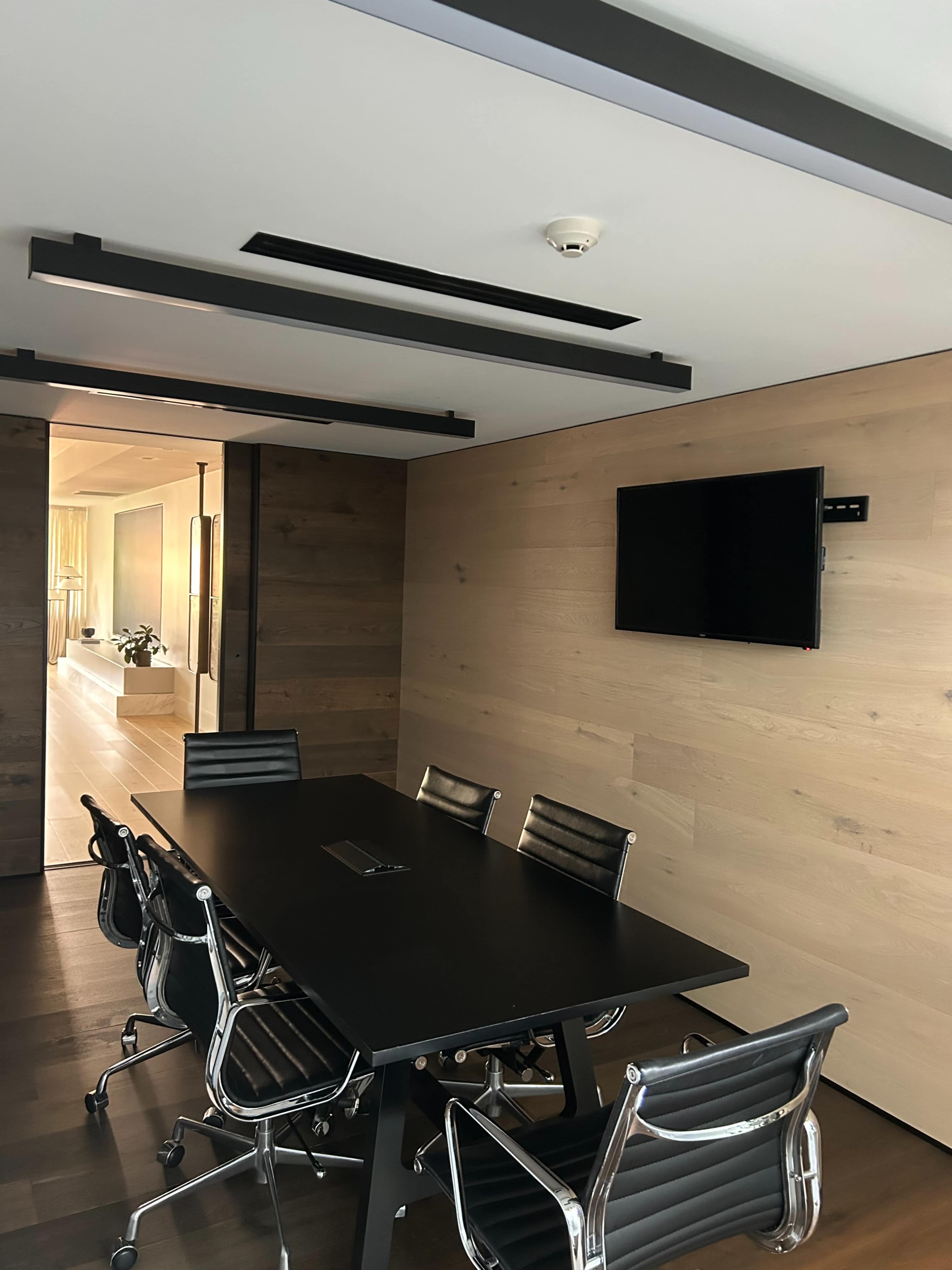 The image shows a modern conference room with a black table, six metal and leather chairs, a flat-screen TV mounted on the wall, and wooden paneling.