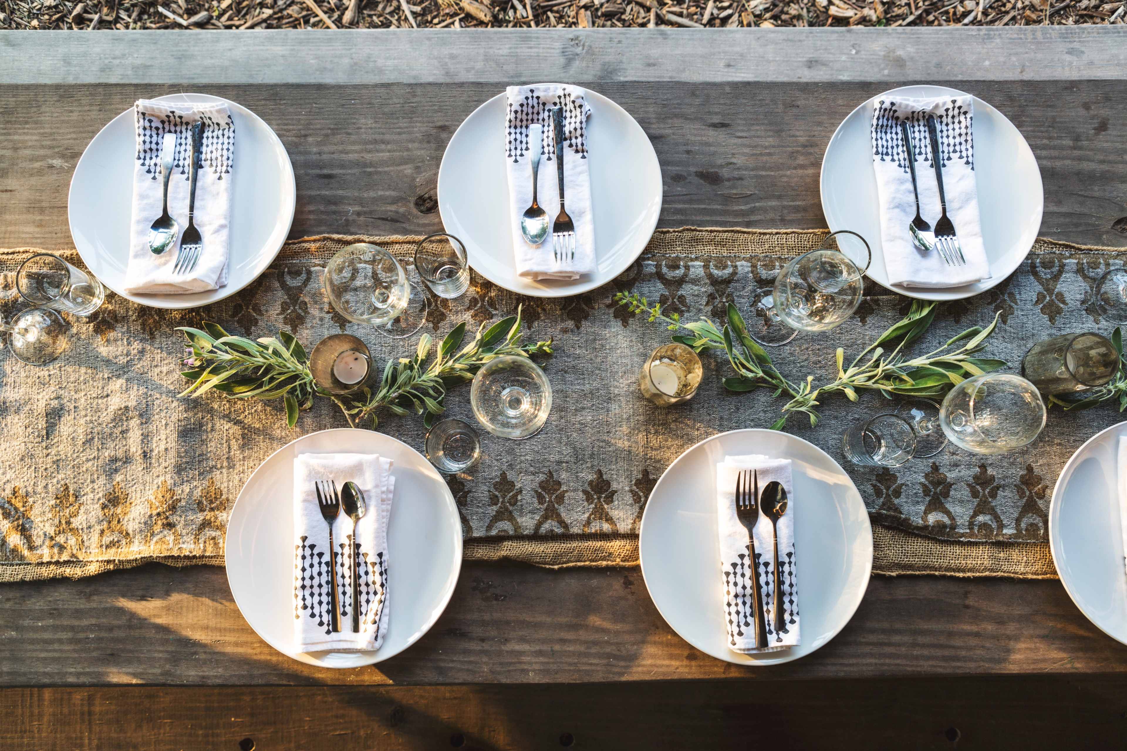 A rectangular table setting displays six plates neatly arranged with cutlery, folded napkins, and decorative glassware, all centered on a textured table runner adorned with greenery.