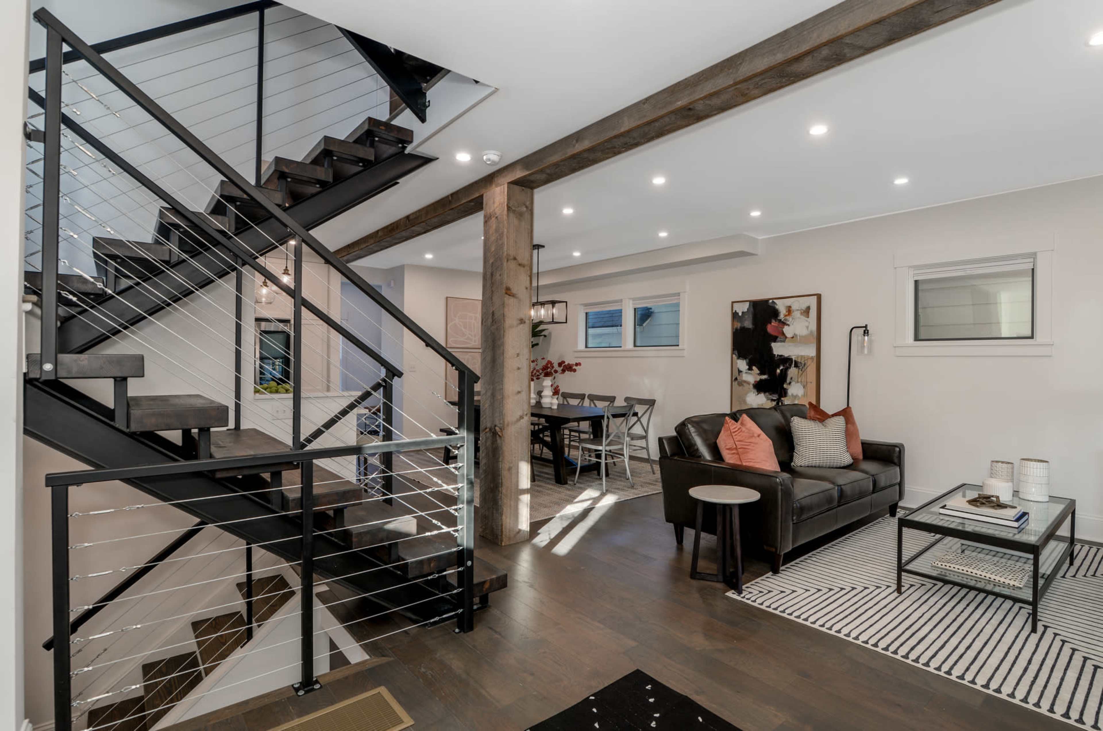 A modern open-concept living area featuring a staircase with metal railings, a seating area with a black sofa, and a dining space in the background.