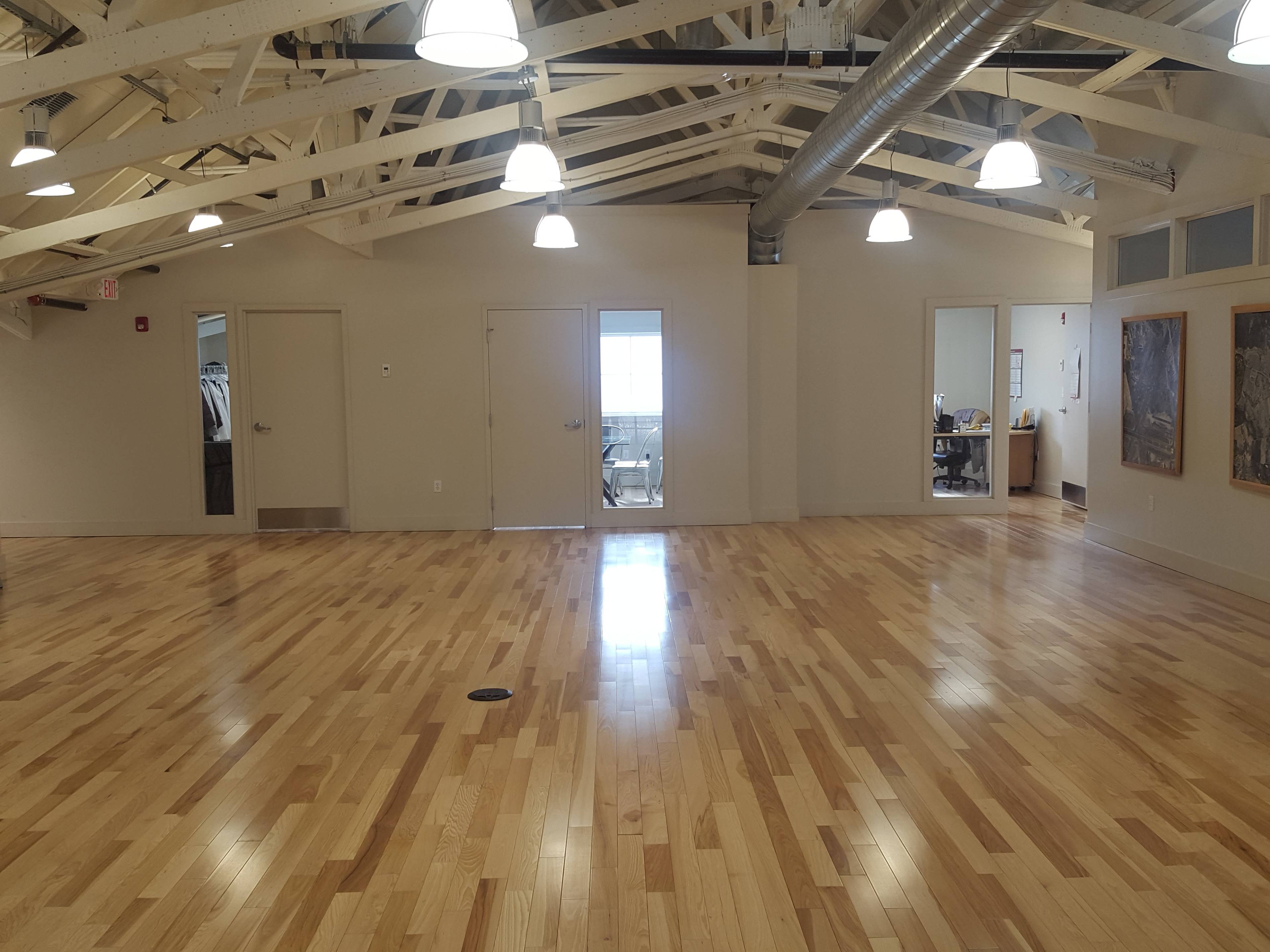 A spacious room with wooden floors, two doors on the left, and large windows illuminating the space.