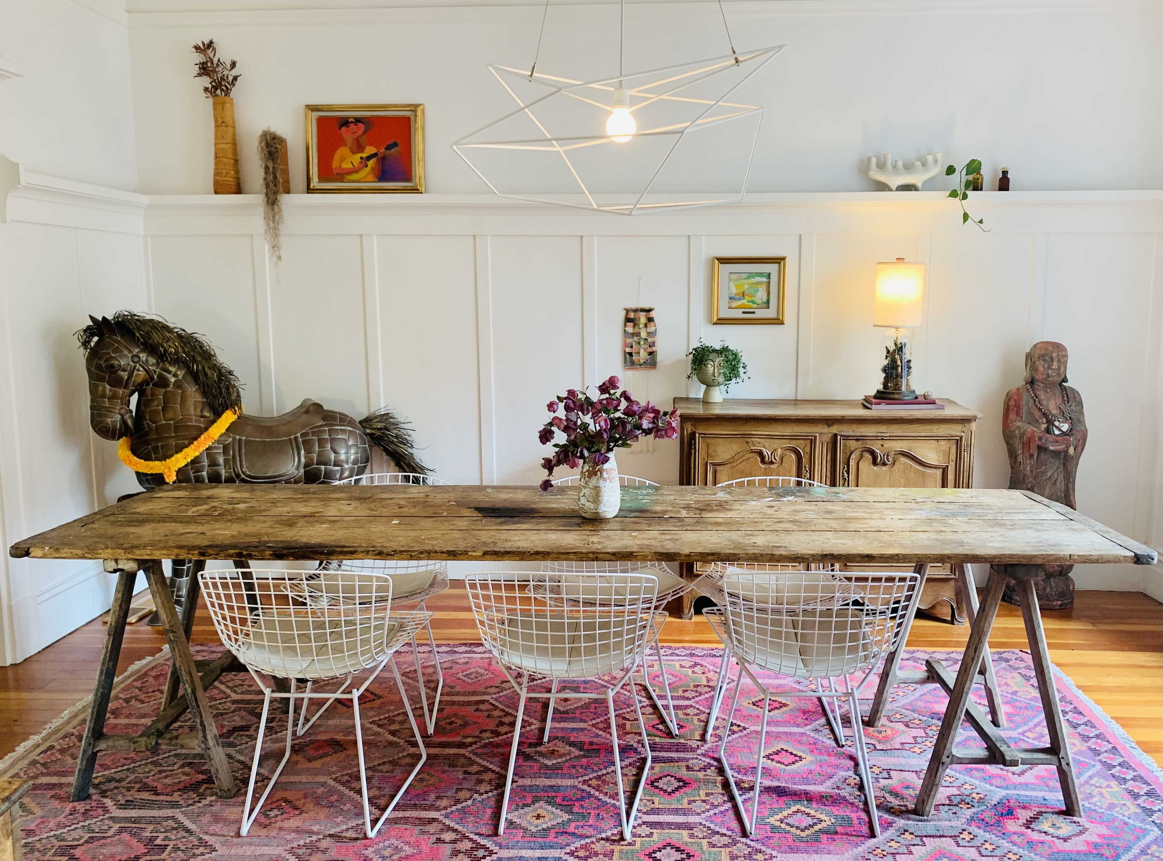 A rustic wooden dining table is surrounded by four white wire chairs in a brightly lit room featuring decorative art pieces, a statue, and an antique sideboard.
