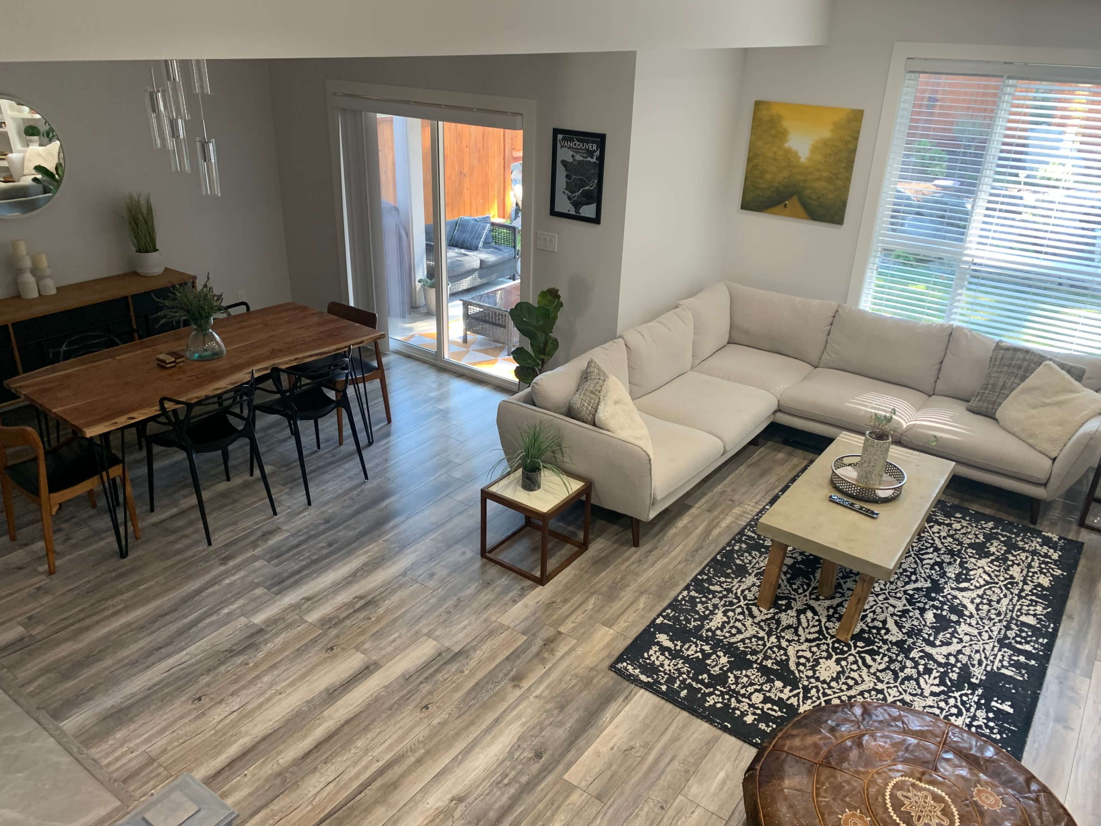 A modern living room and dining area with a light-colored sectional sofa, a wooden dining table, and large windows letting in natural light.