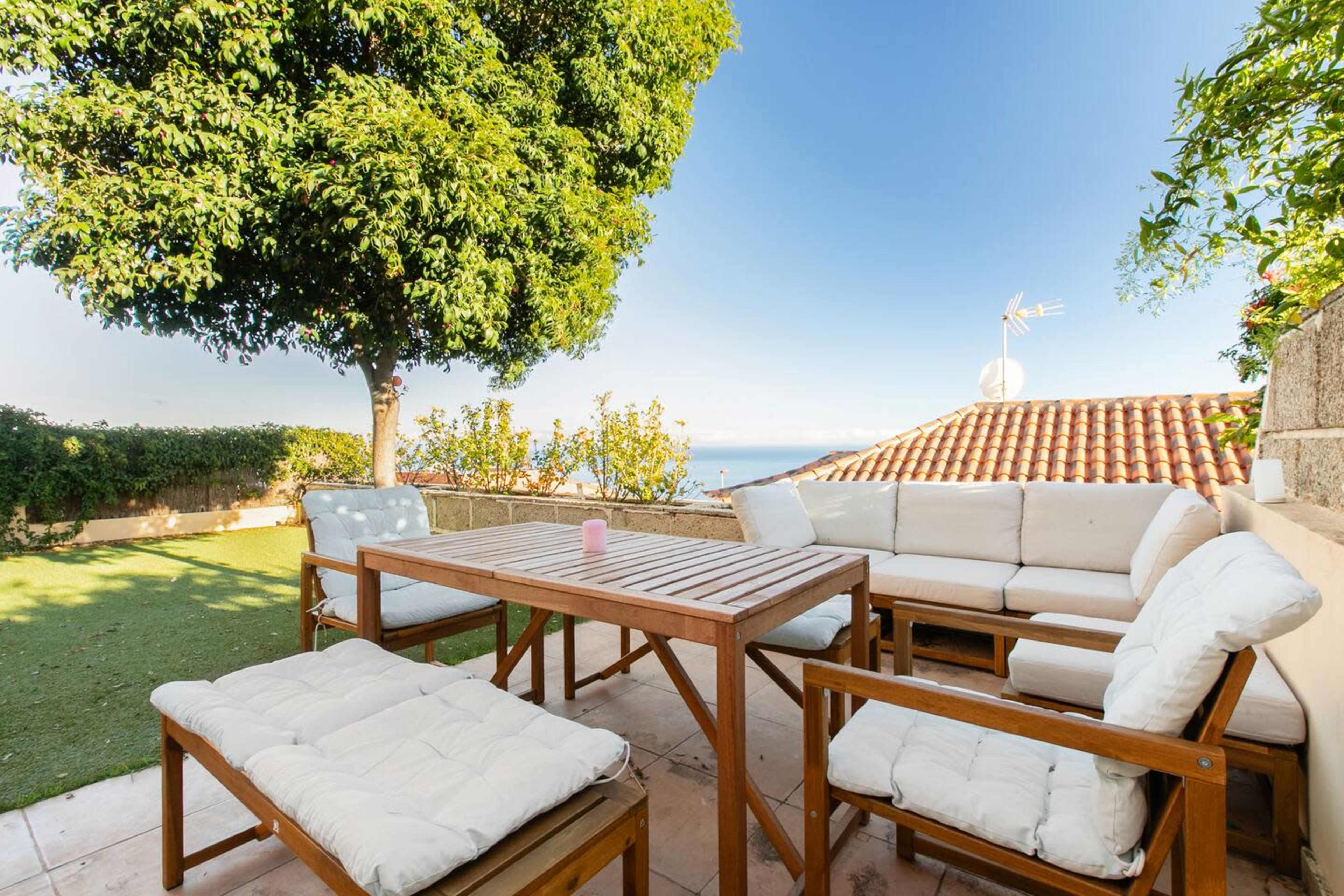 A patio with wooden furniture and cushions overlooks a green lawn and a distant view of the ocean under a clear sky.