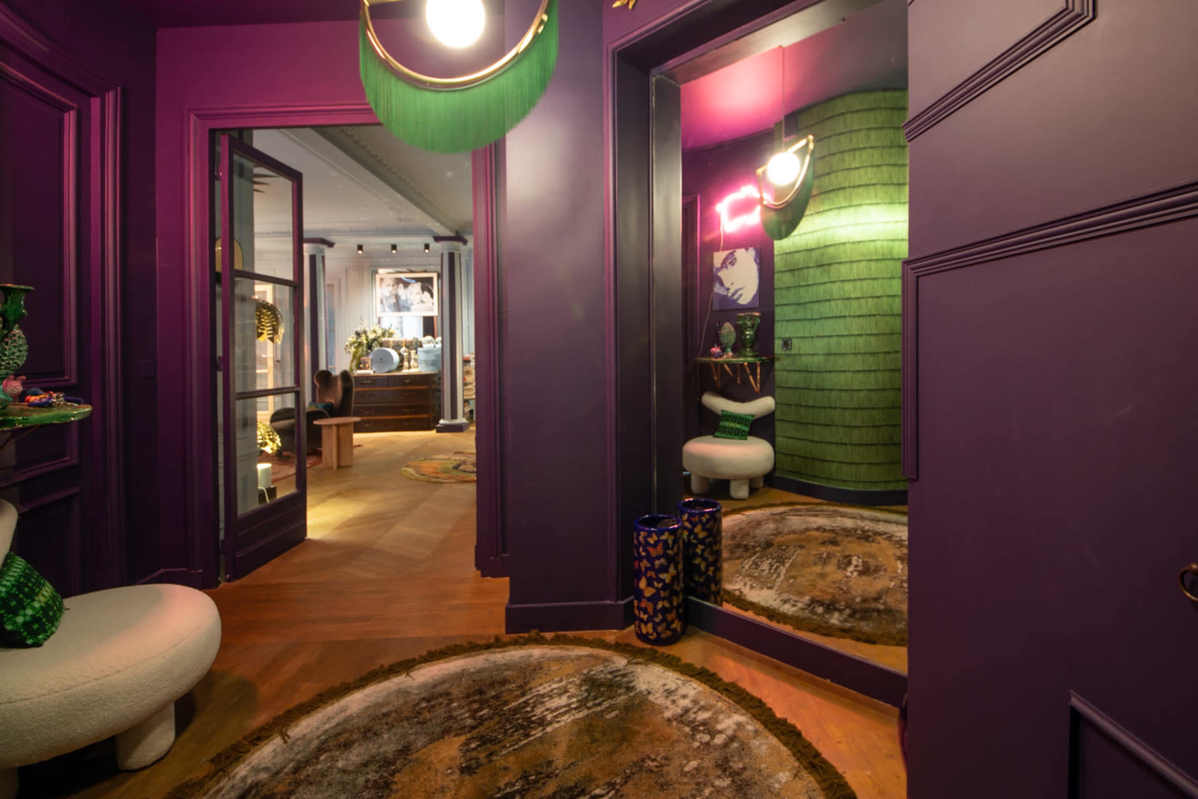The image shows a brightly colored hallway with purple walls, featuring a round rug on the floor, a curved white chair, and a mirrored wall reflecting green decor.