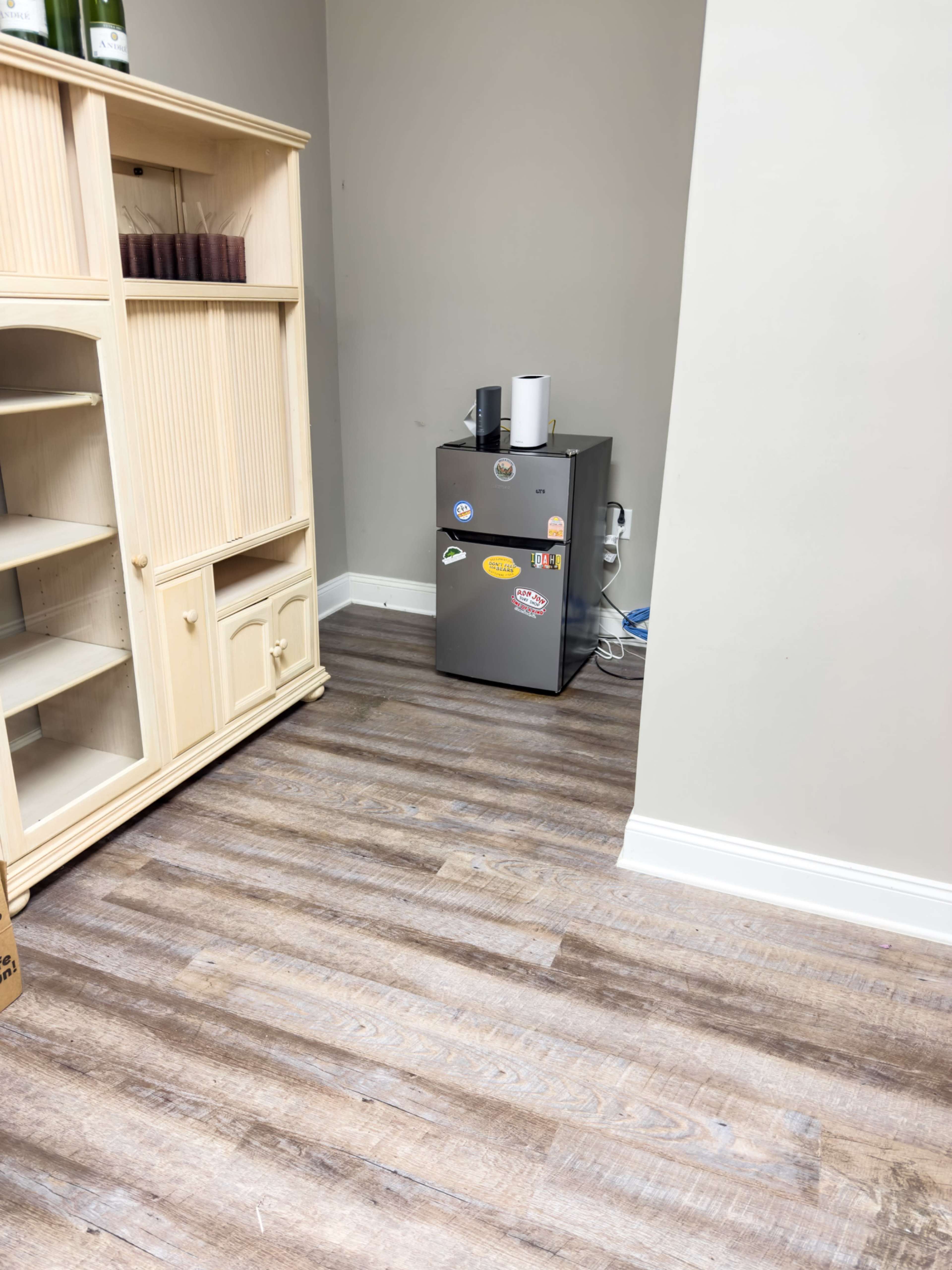 A small corner of a room features a gray refrigerator adorned with various stickers, positioned next to a light-colored wooden shelving unit on a wooden floor.