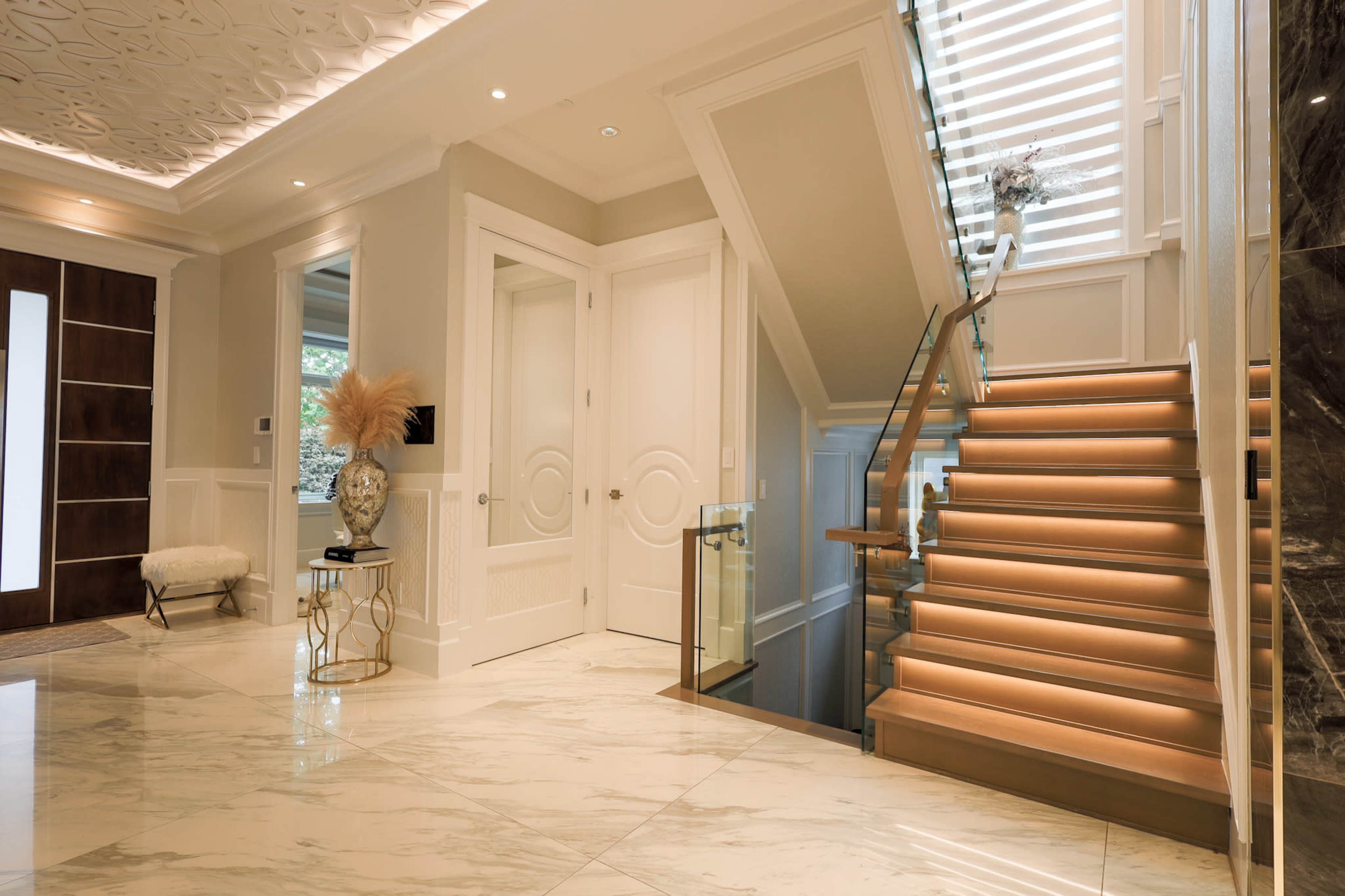 The image features a spacious foyer with marble flooring, a staircase with wooden steps and glass railings, and elegant lighting.